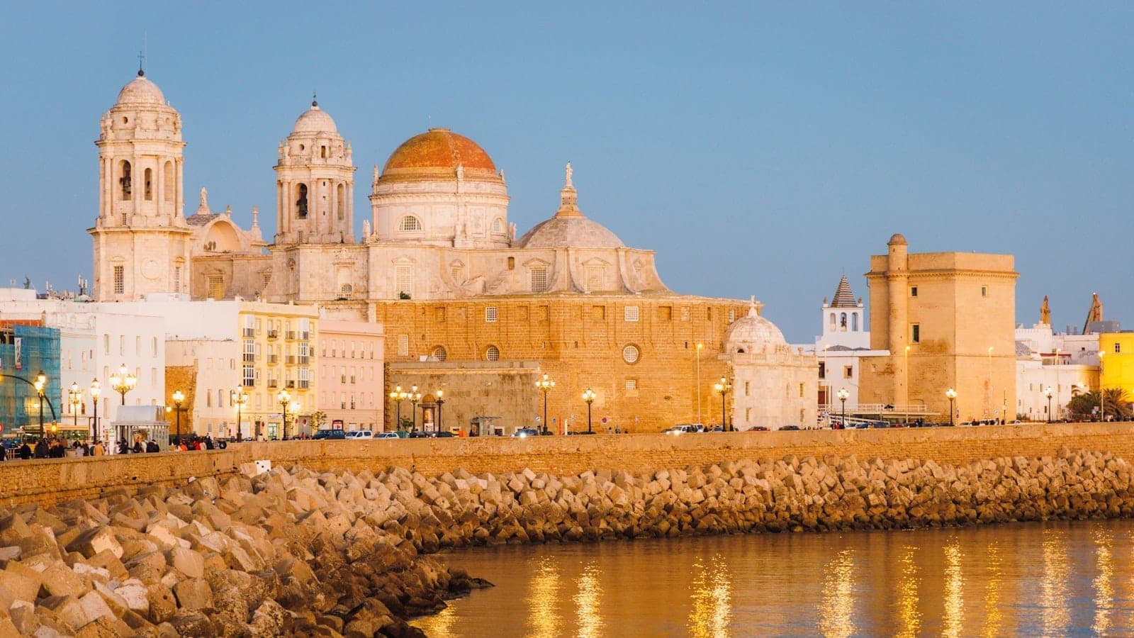 Tanawing ginintuan ng araw ng Baroque na katedral ng Cádiz at mga makasaysayang gusali sa tabing-dagat na aninag sa tubig, may lumang pader ng lungsod at maliwanag na bughaw na langit.