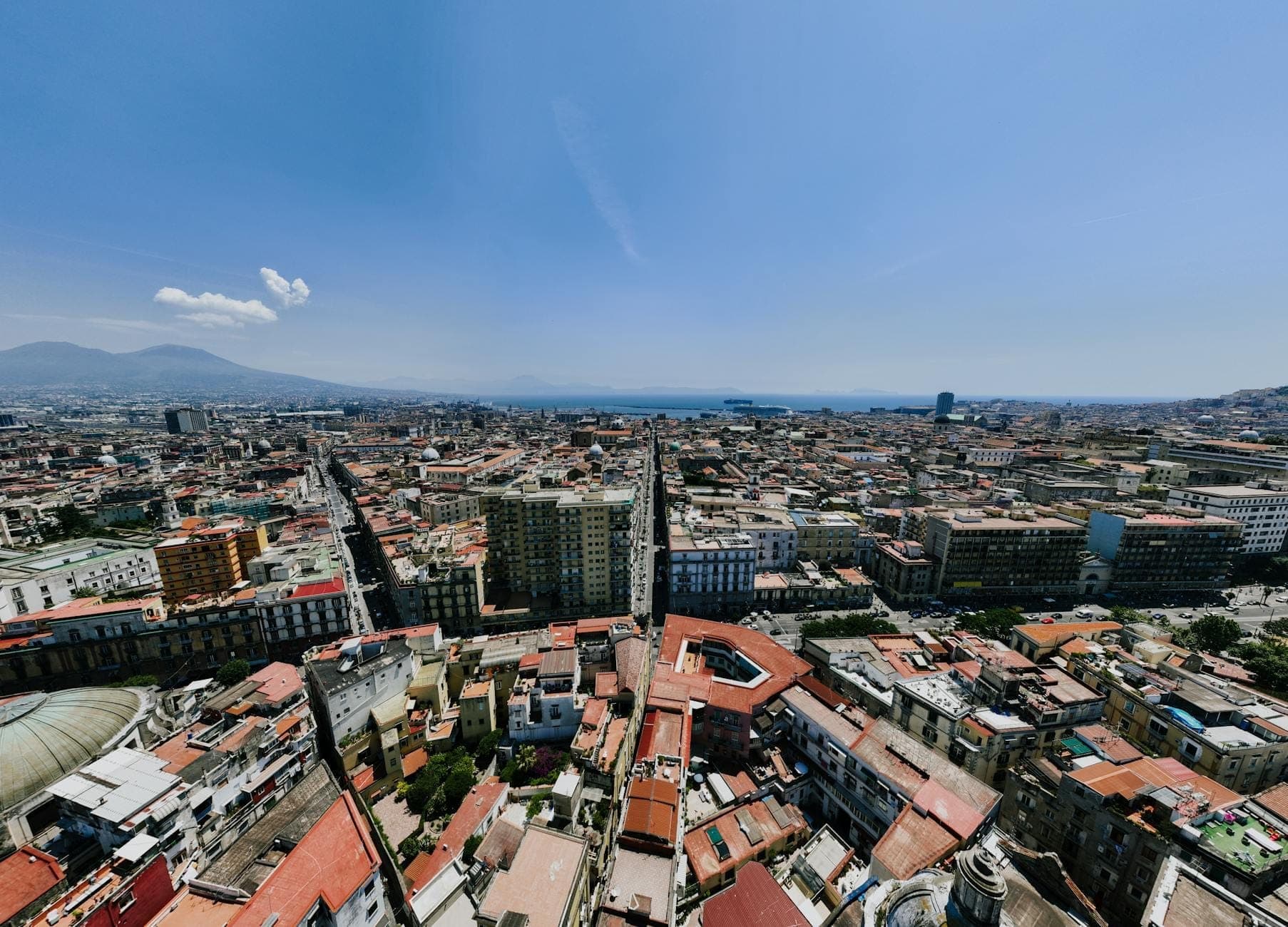 Polong-tingin ng Naples Centro Storico na nagpapakita ng makapal na makasaysayang mga gusali, tuwid na mga kalye ng decumanus, at malalayong look sa ilalim ng maliwanag na asul na langit.