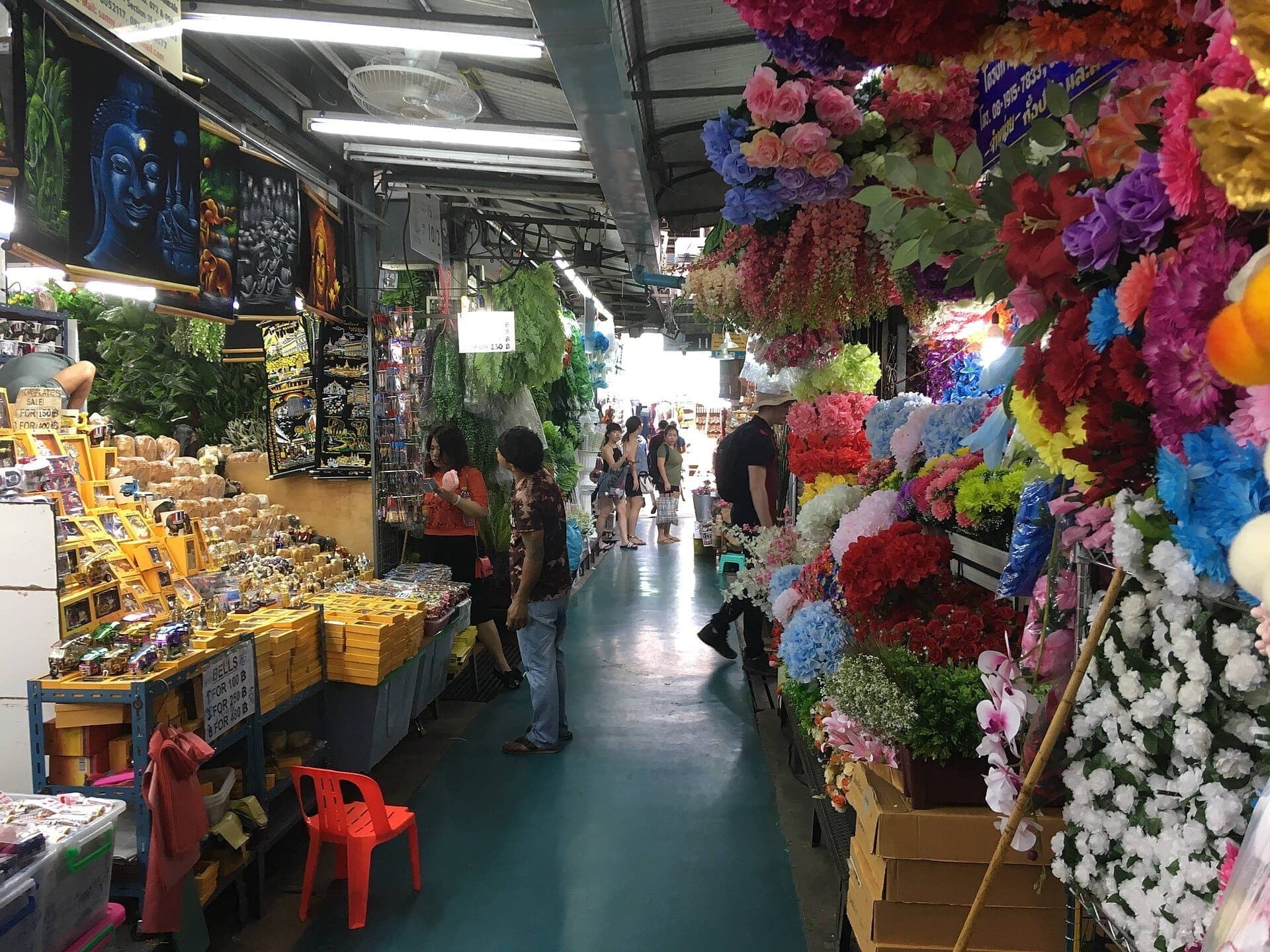Lối đi bên trong chợ cuối tuần Chatuchak với hoa giả sặc sỡ, gian hàng quà lưu niệm và du khách đang dạo mua sắm ở Bangkok