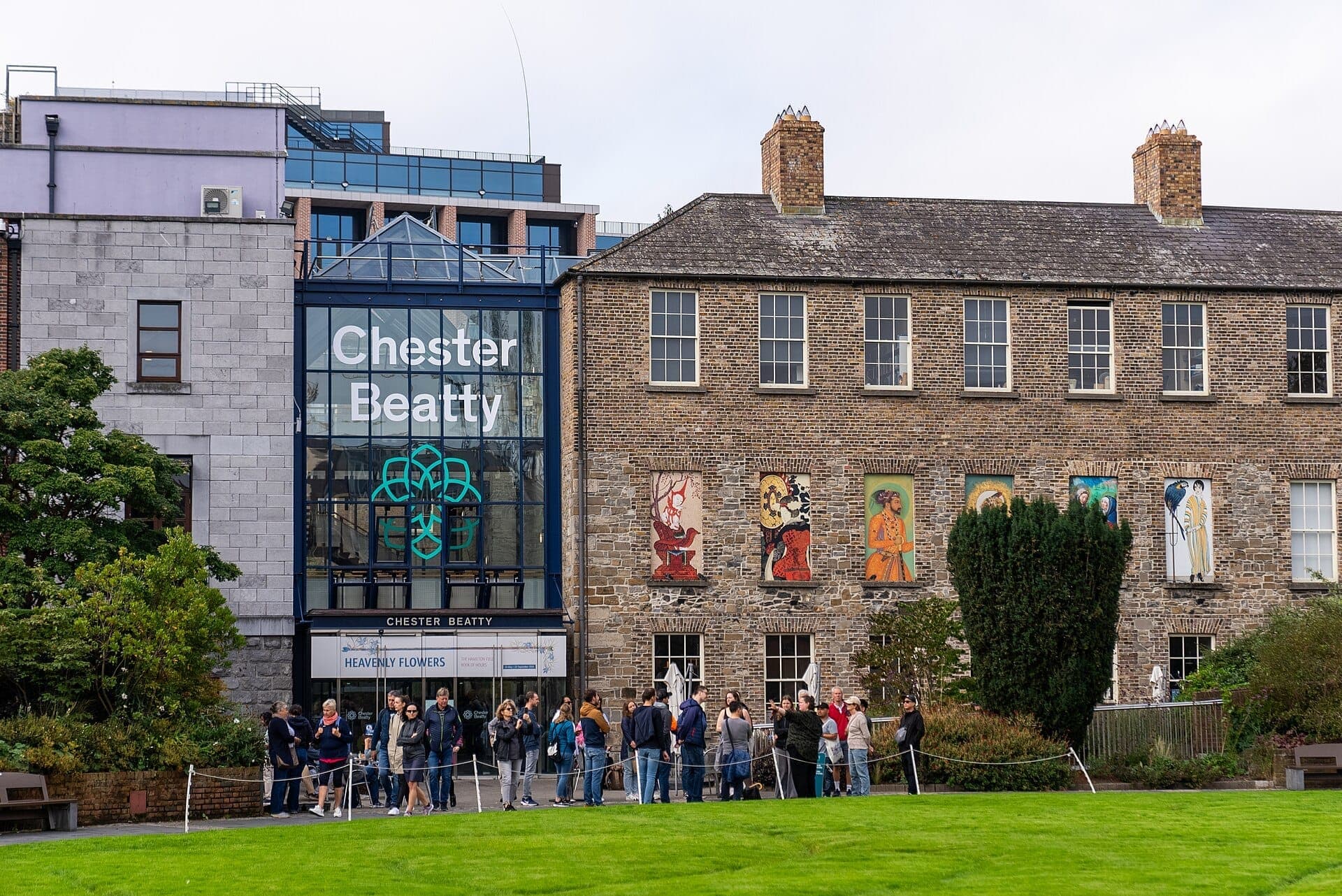 Vista exterior de la Chester Beatty Library en Dublín con entrada de cristal moderna, muros de ladrillo históricos, paneles de arte coloridos y visitantes esperando fuera.