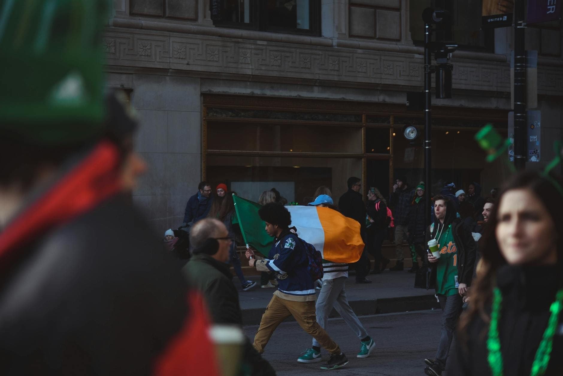Menschenmenge feiert St. Patrick's Day im Freien, mit einem Mann, der eine irische Flagge eine Stadtstraße entlangträgt, Gebäude im Hintergrund.