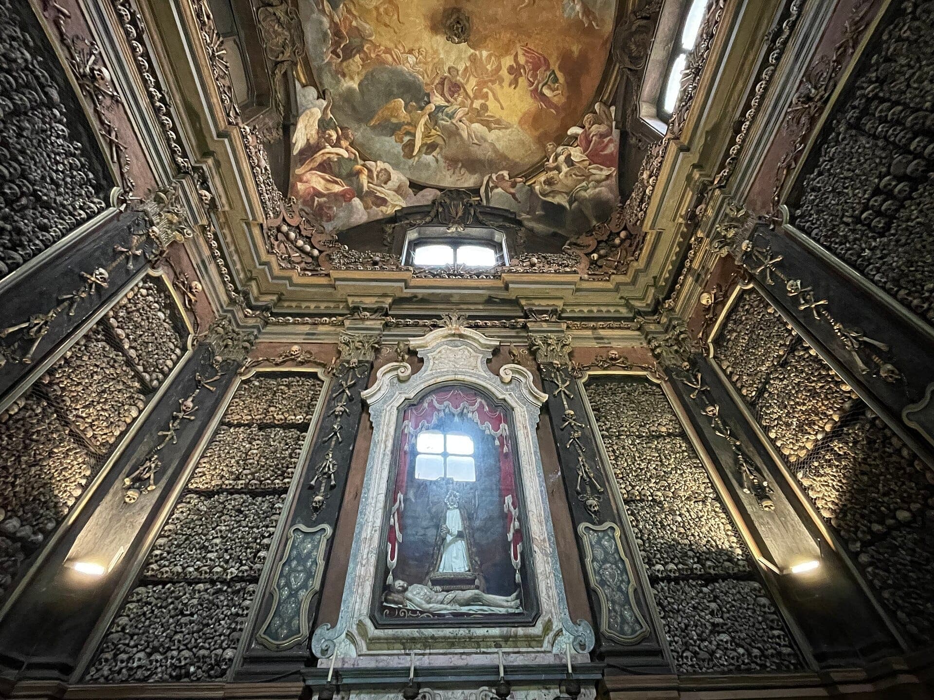 Interior of the Chiesa di San Bernardino alle Ossa ossuary chapel, showing walls covered in human skulls and bones, with a painted baroque ceiling.
