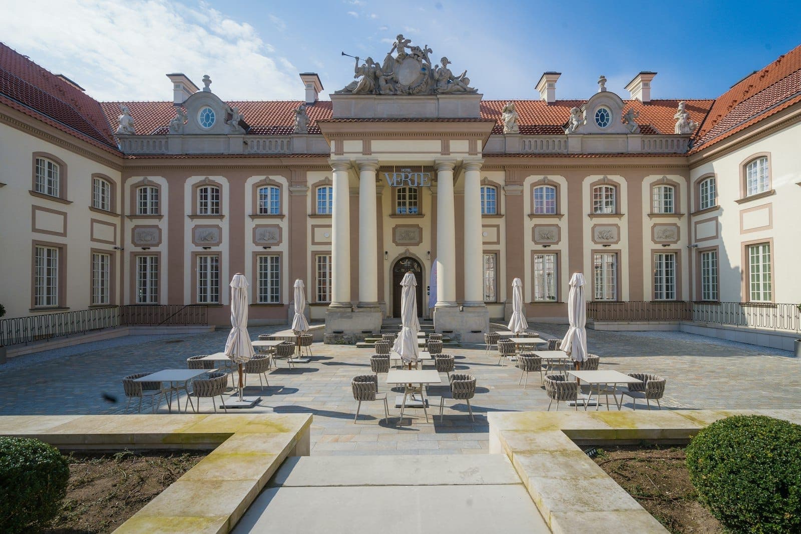 Elegante cortile di un palazzo barocco con tavolini da caffè, ombrelloni e sculture ornate sopra l'ingresso, sotto un cielo blu a Varsavia.