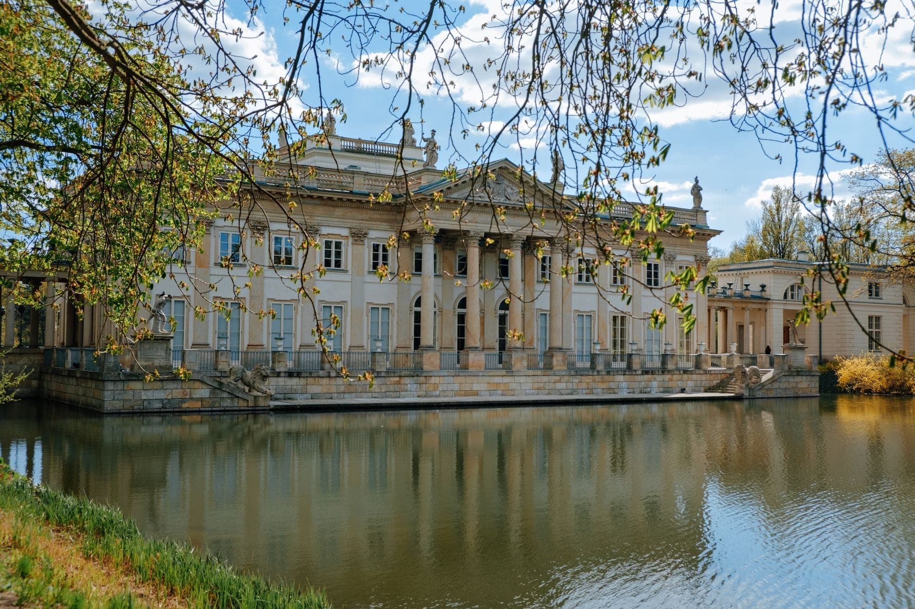 Neoklassizistischer Palast an einem ruhigen Teich im Warschauer Łazienki-Park, eingerahmt von Ästen und blauem Himmel – ein Ort, der Musikliebhaber in Chopins Welt einlädt.