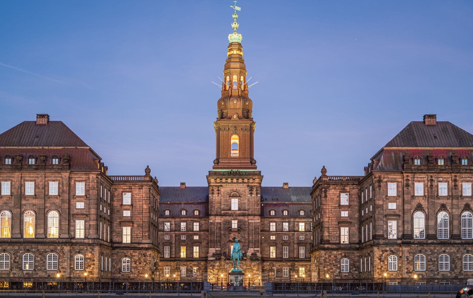 Schloss Christiansborg in der Abenddämmerung mit seinem beleuchteten Mittelturm und einer Statue davor, von einem weiten freien Platz unter einem klaren blauen Abendhimmel aus gesehen.