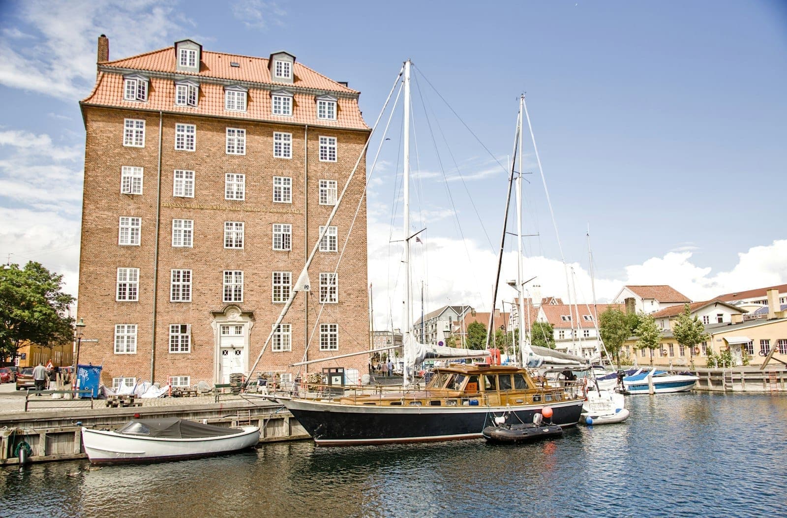 Ein historisches Backsteingebäude mit roten Dachziegeln säumt den Christianshavn-Kanal, mit Segelbooten und kleinen Booten bei sonnigem Wetter vor Anker.