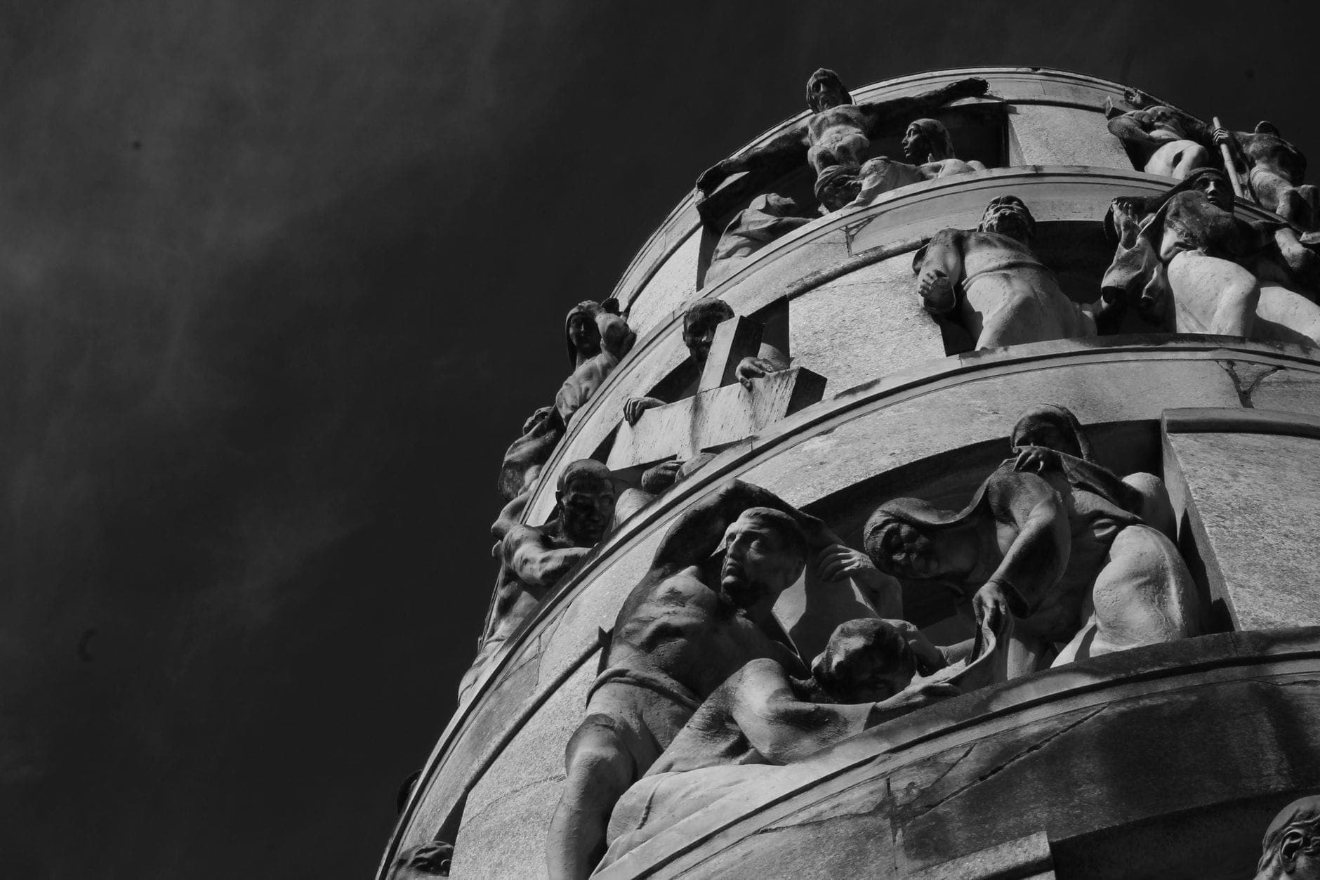 Striking black and white view looking up at a large round funeral monument adorned with dynamic human sculptures against the sky in Cimitero Monumentale di Milano.