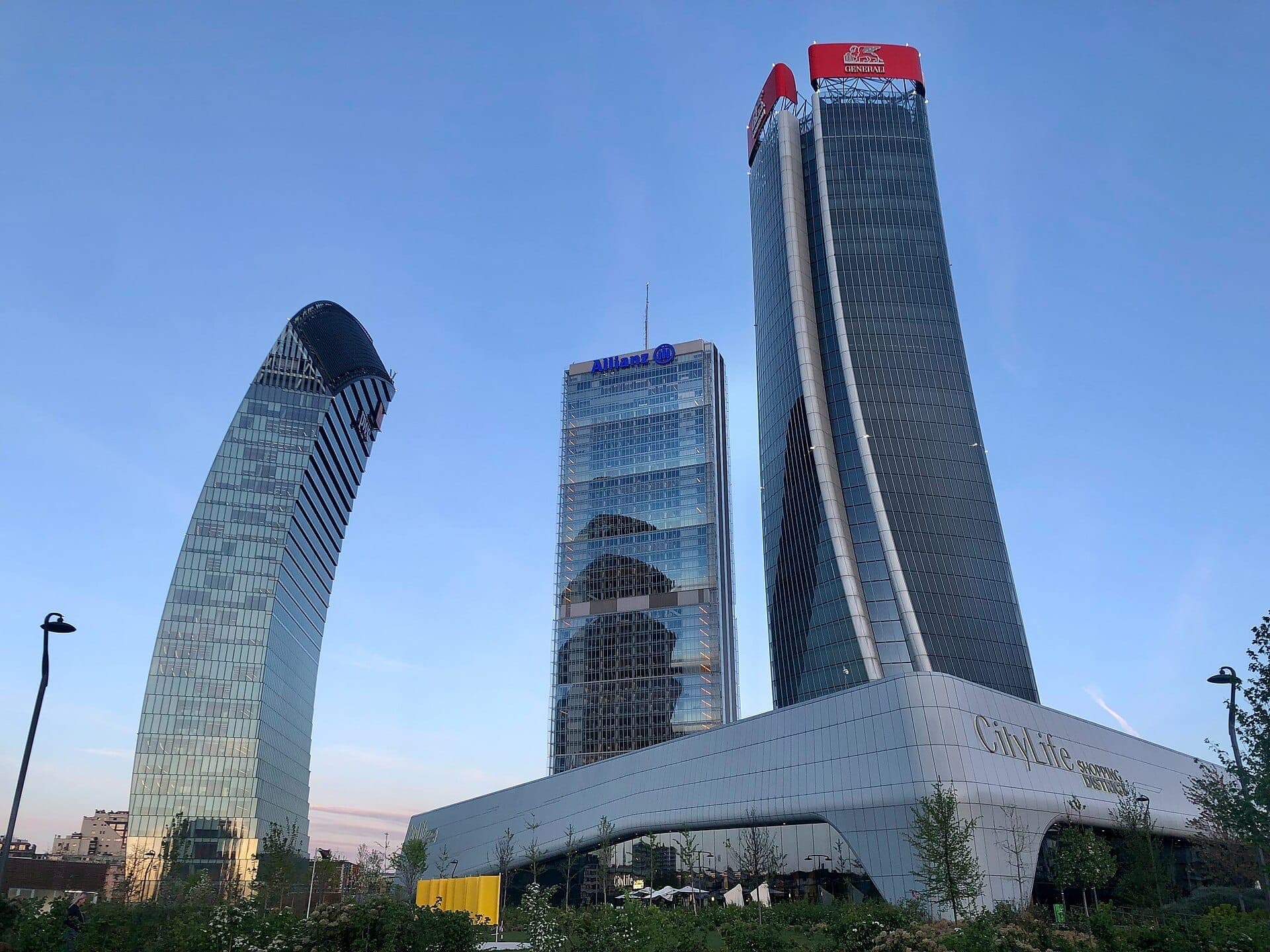 Three modern skyscrapers at CityLife in Milan rise above a green park under a clear blue sky at sunset.