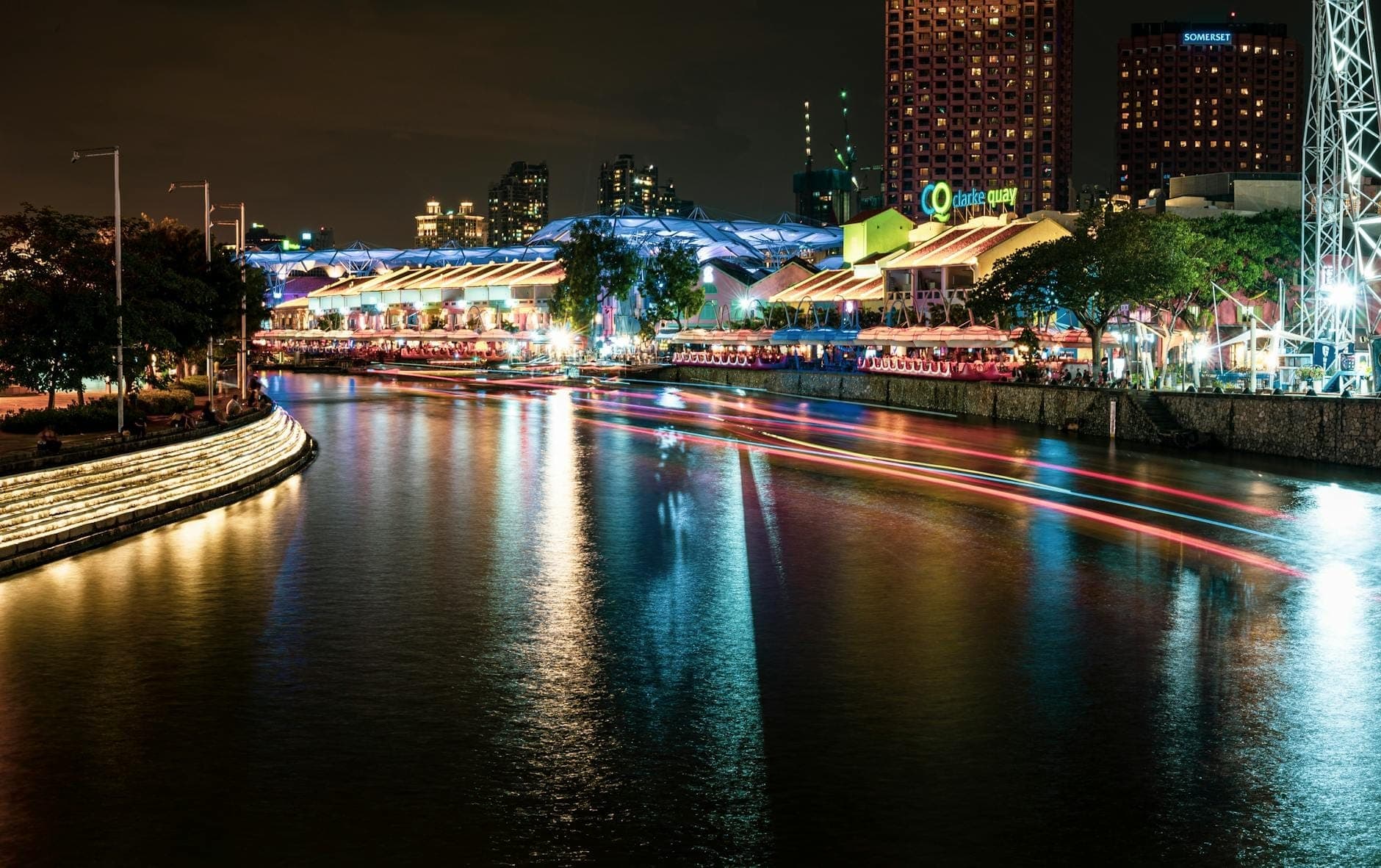Night view of Clarke Quay with colorful lights reflecting on the Singapore River, bustling riverside shophouses, and vibrant nightlife atmosphere.