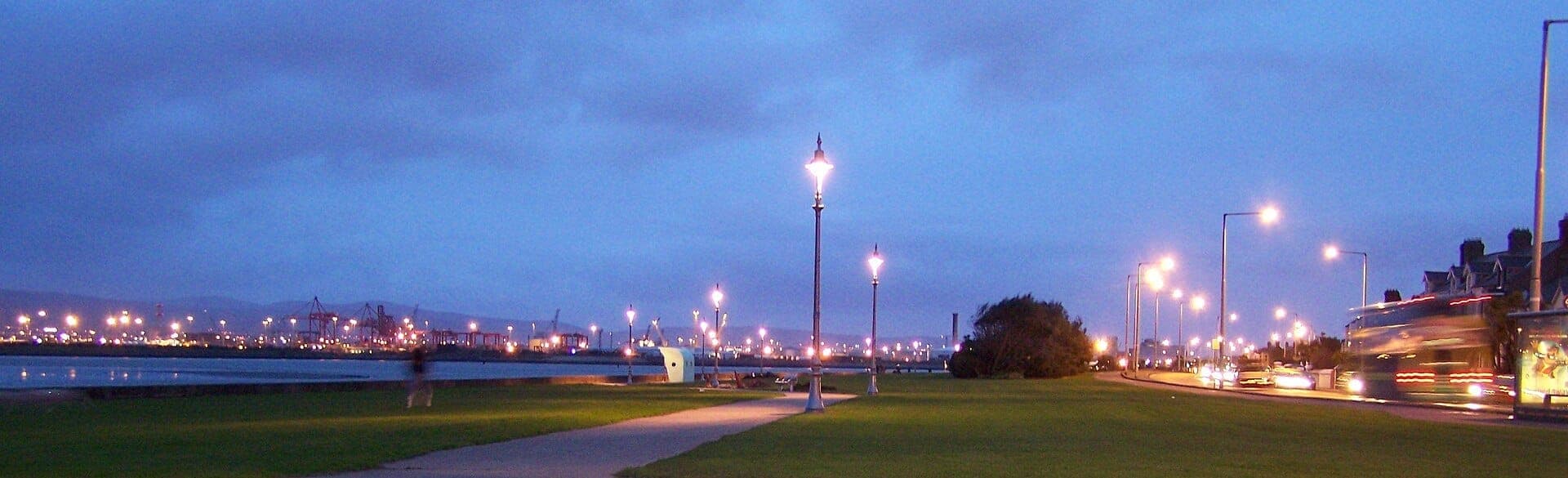 Clontarf Promenade in der Dämmerung mit beleuchteten Straßenlaternen, einem Grünstreifen am Meer, Stadtlichtern und vorbeifahrendem Verkehr unter blauem Himmel.