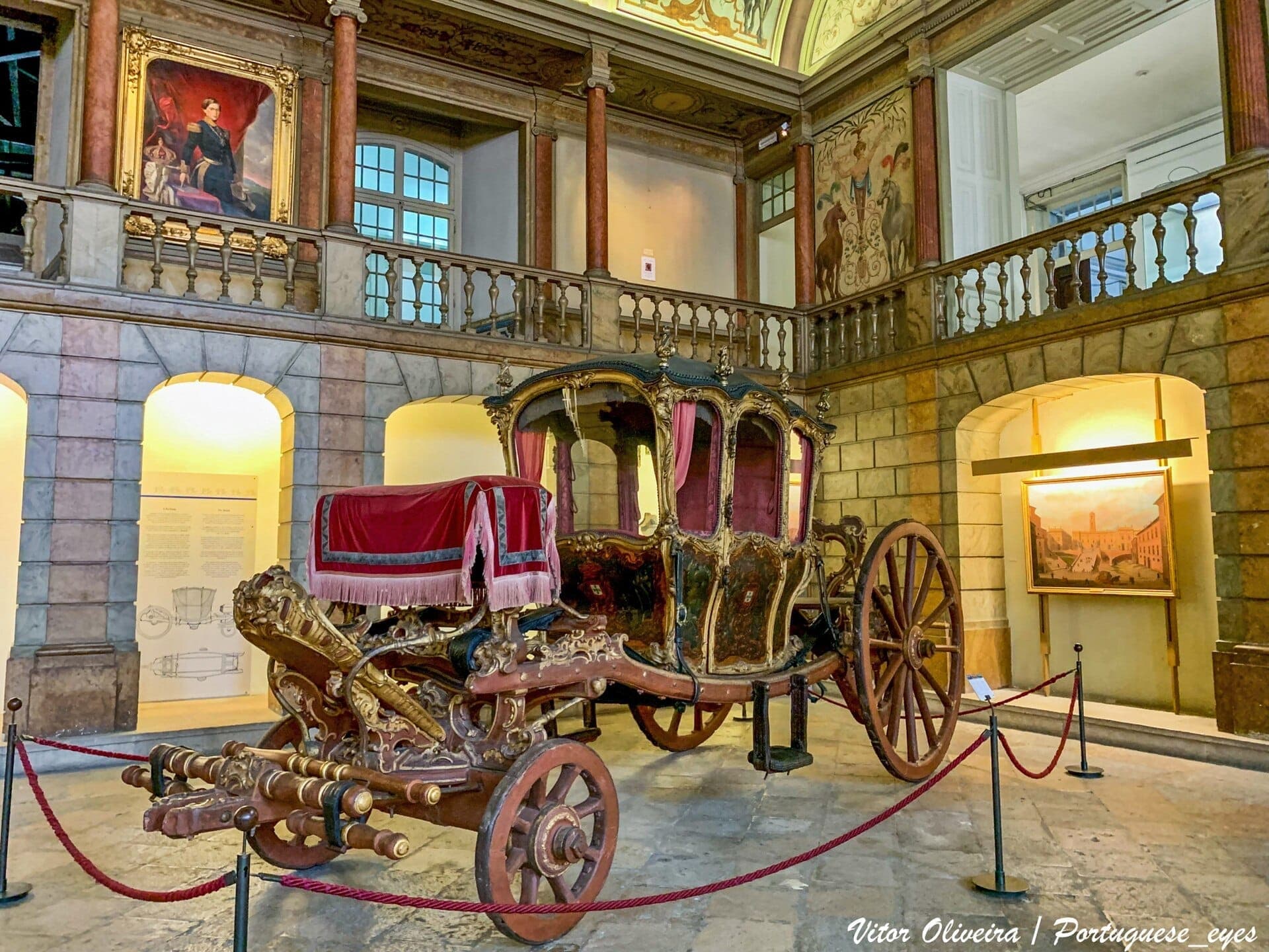 Cỗ xe ngựa hoàng gia được trang trí lộng lẫy trưng bày bên trong Museu Nacional dos Coches tại Lisbon, bao quanh bởi các bức tranh lịch sử, cột đá cẩm thạch và ánh đèn bảo tàng dịu nhẹ.