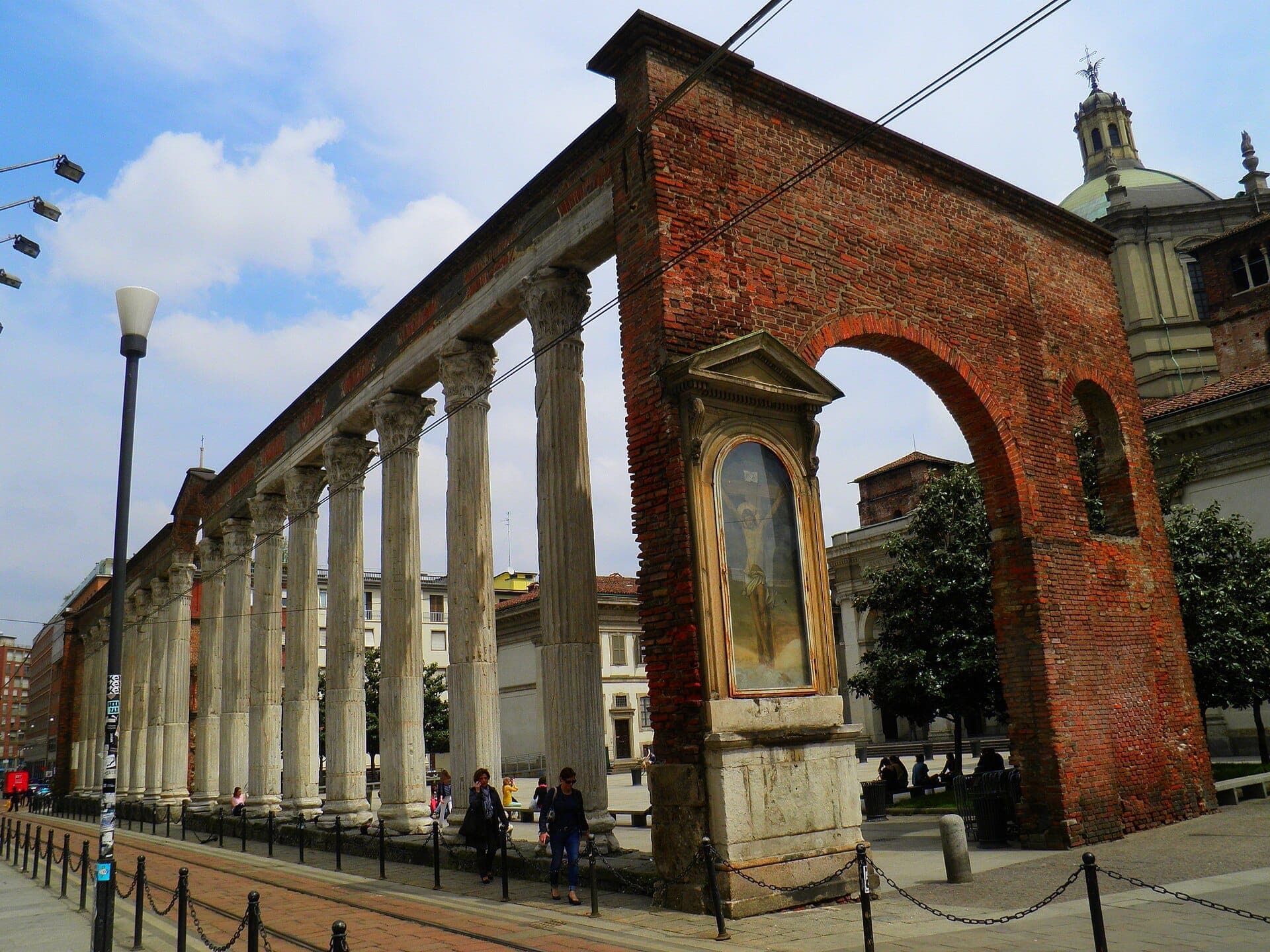 The Colonne di San Lorenzo in Milan, featuring sixteen ancient marble Corinthian columns and red brick arches, under a partly cloudy sky.