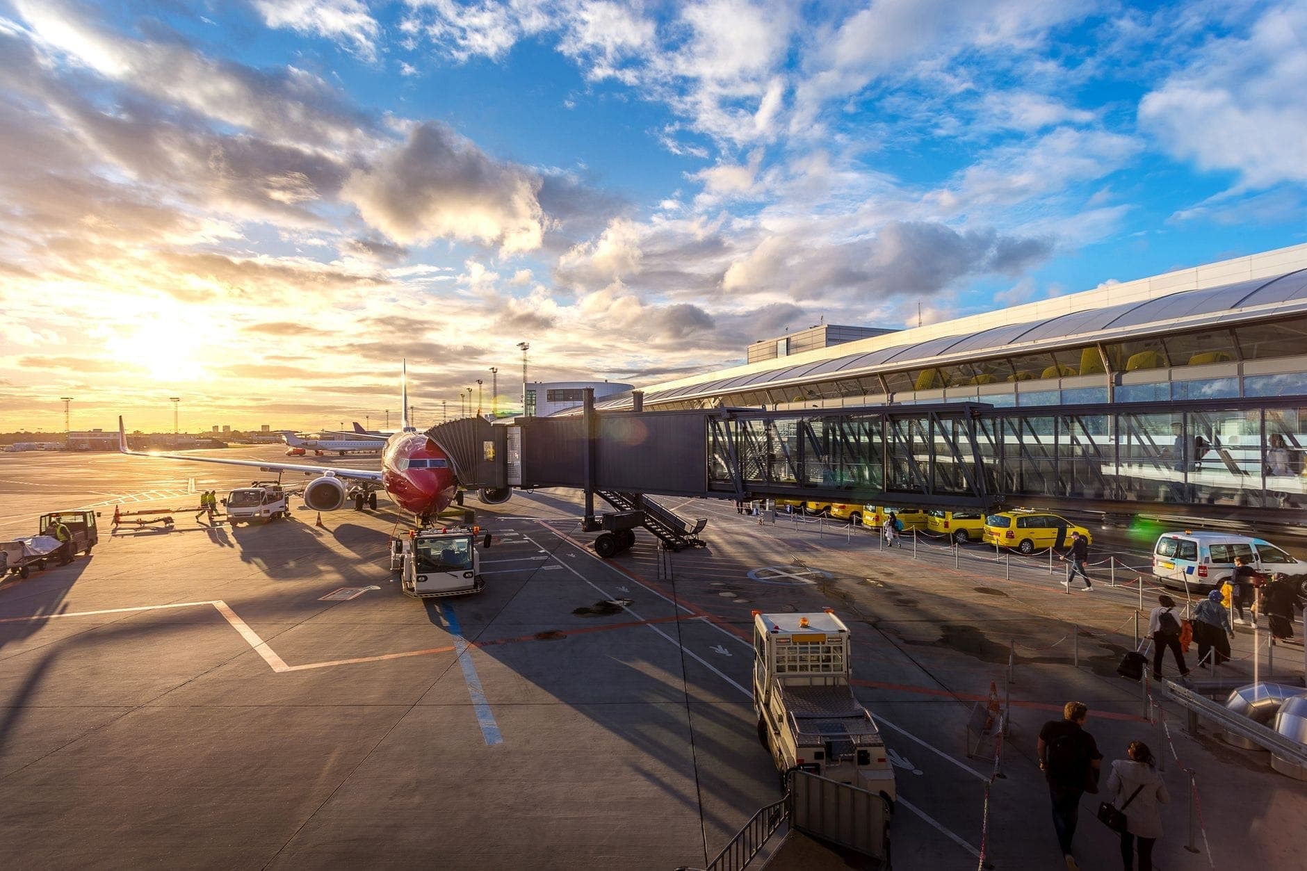 Der Kopenhagener Flughafen bei Sonnenaufgang mit einem Flugzeug am Gate, gelben Taxis, dem Terminalgebäude und Passagieren auf dem Rollfeld unter einem leuchtenden Himmel.