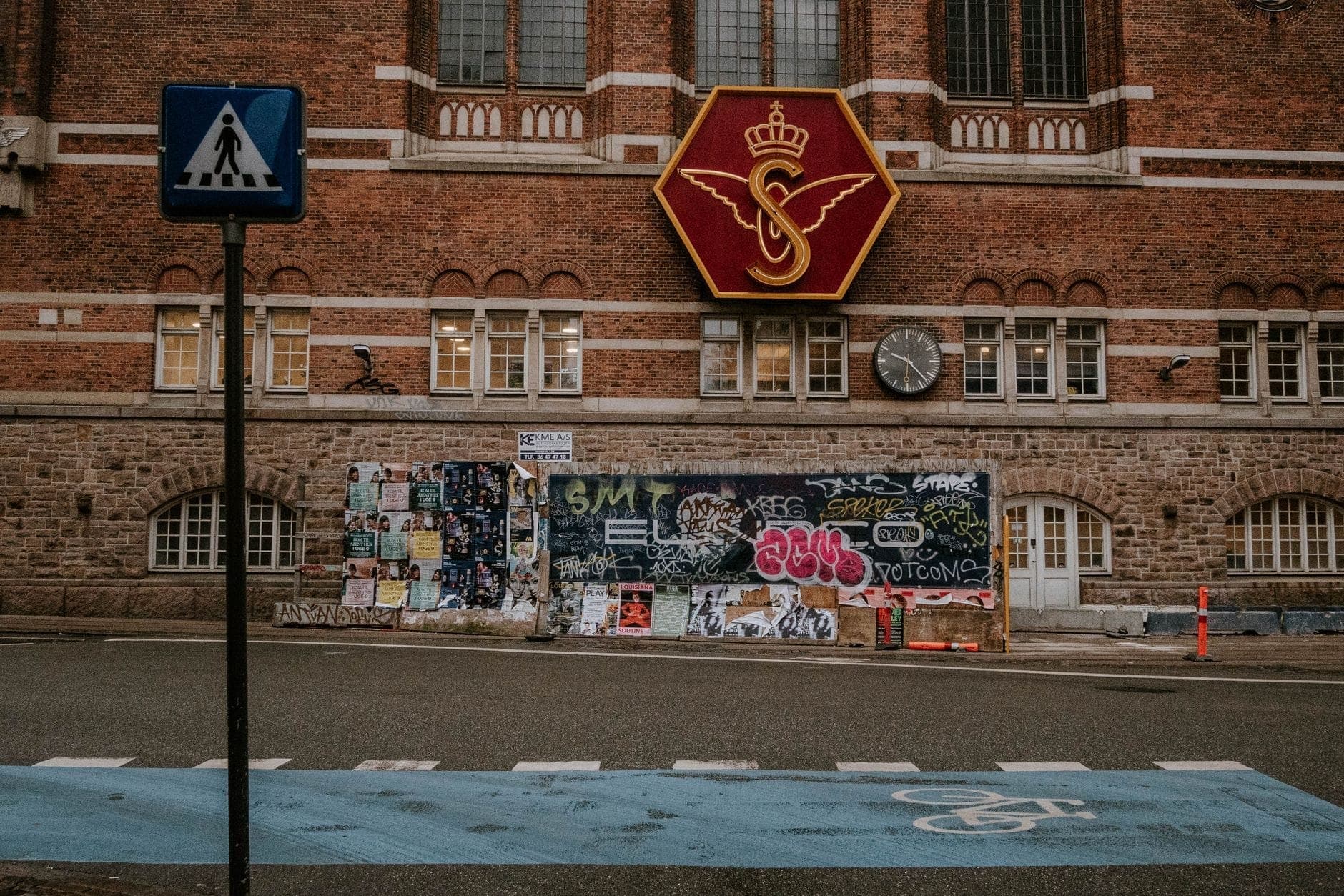 Widok z poziomu ulicy na historyczny ceglany budynek stacji w Kopenhadze z dużym szyldem skrzydlatego koła DSB i zegarem na fasadzie.