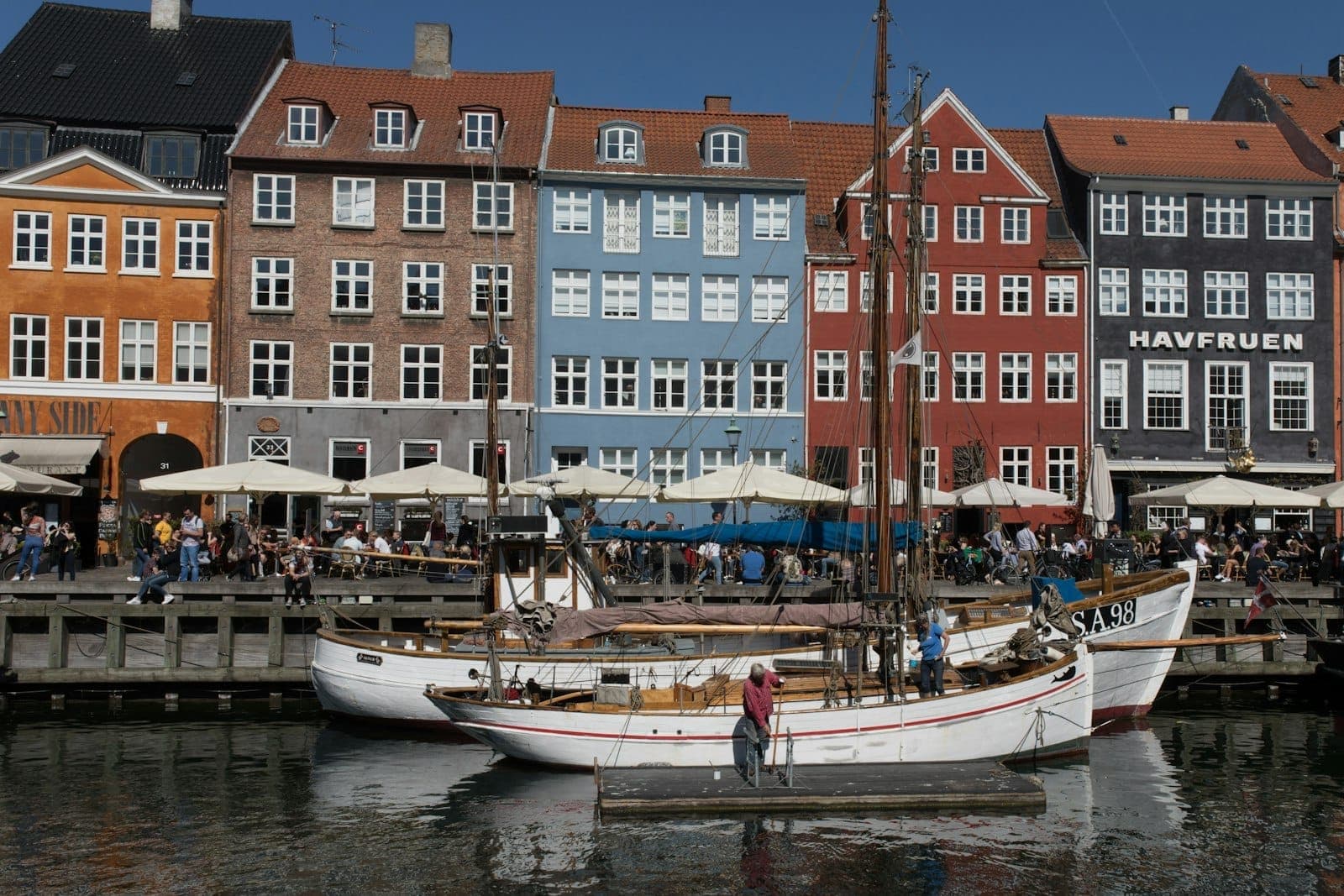 Bunte historische Häuser und Boote am Nyhavn in Kopenhagen an einem sonnigen Tag, mit Menschen in Cafés und beim Sightseeing.
