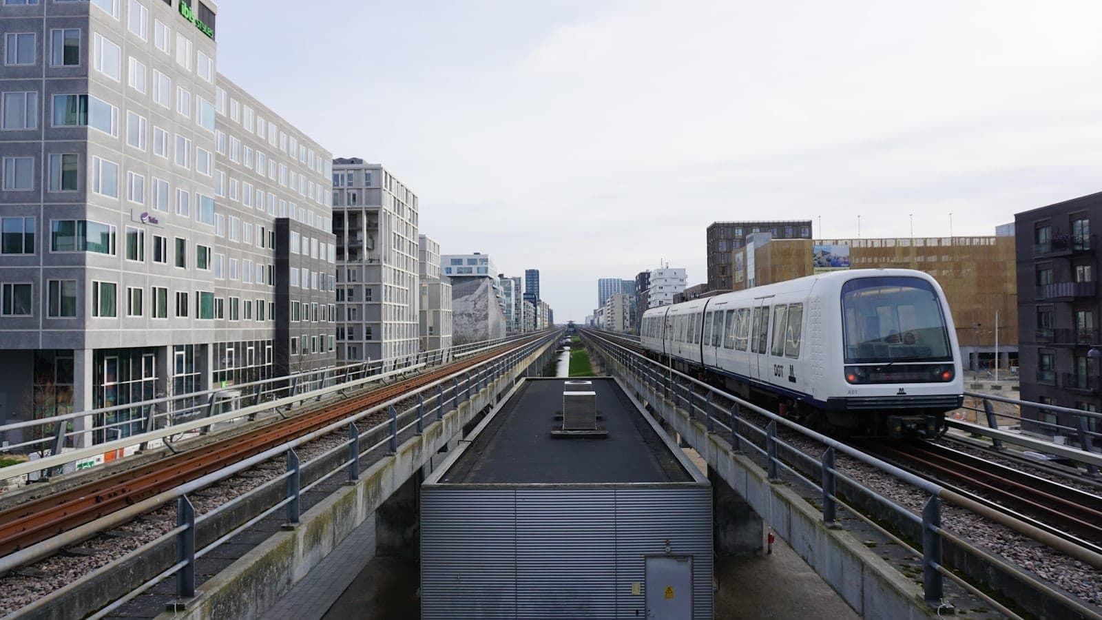 Pociąg metra Copenhagen jadący po estakadzie z nowoczesnymi budynkami miejskimi po obu stronach, pod zachmurzonym niebem.