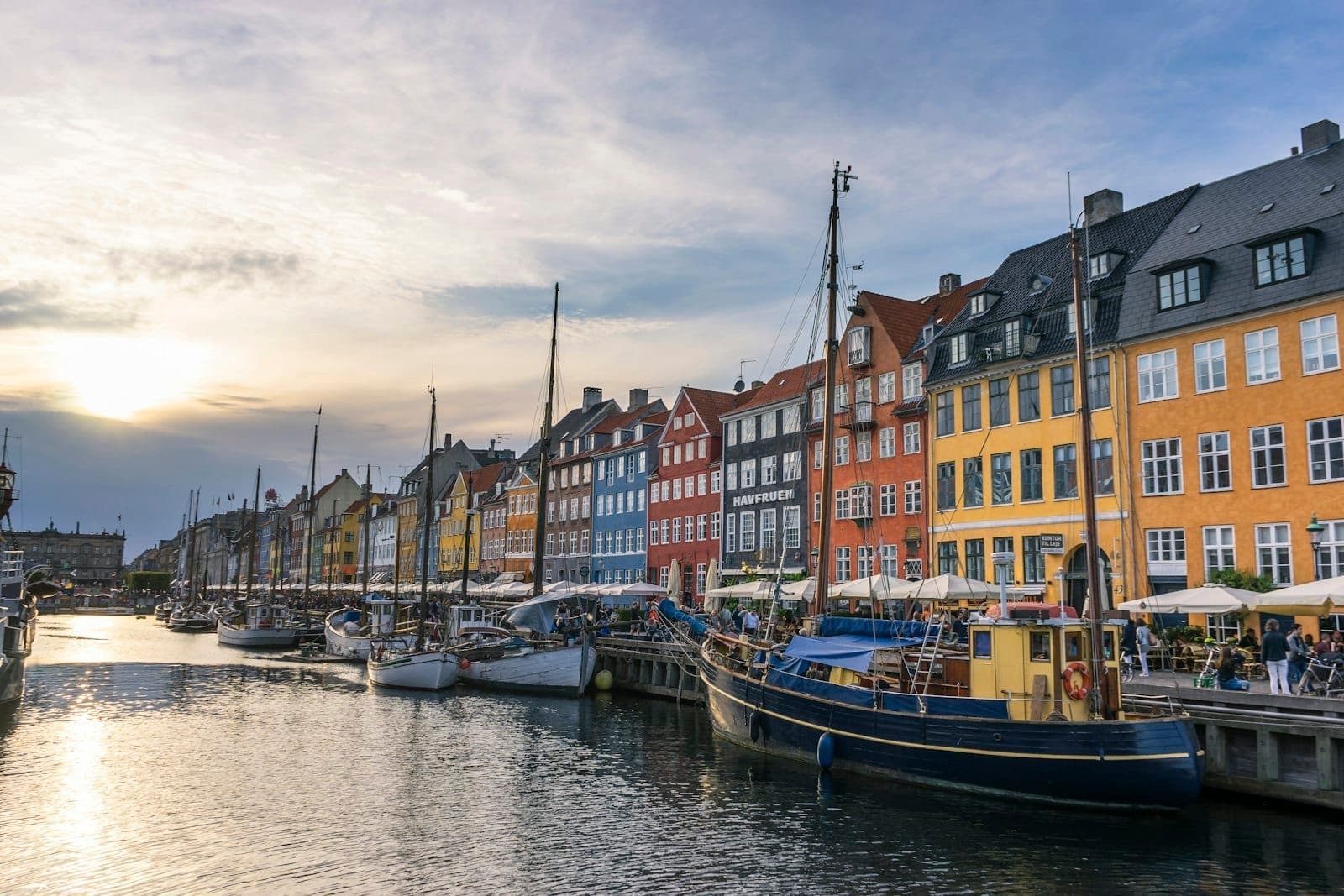 Colorful historic buildings and boats along Nyhavn canal in Copenhagen at sunset, with blue sky and lively waterfront atmosphere.