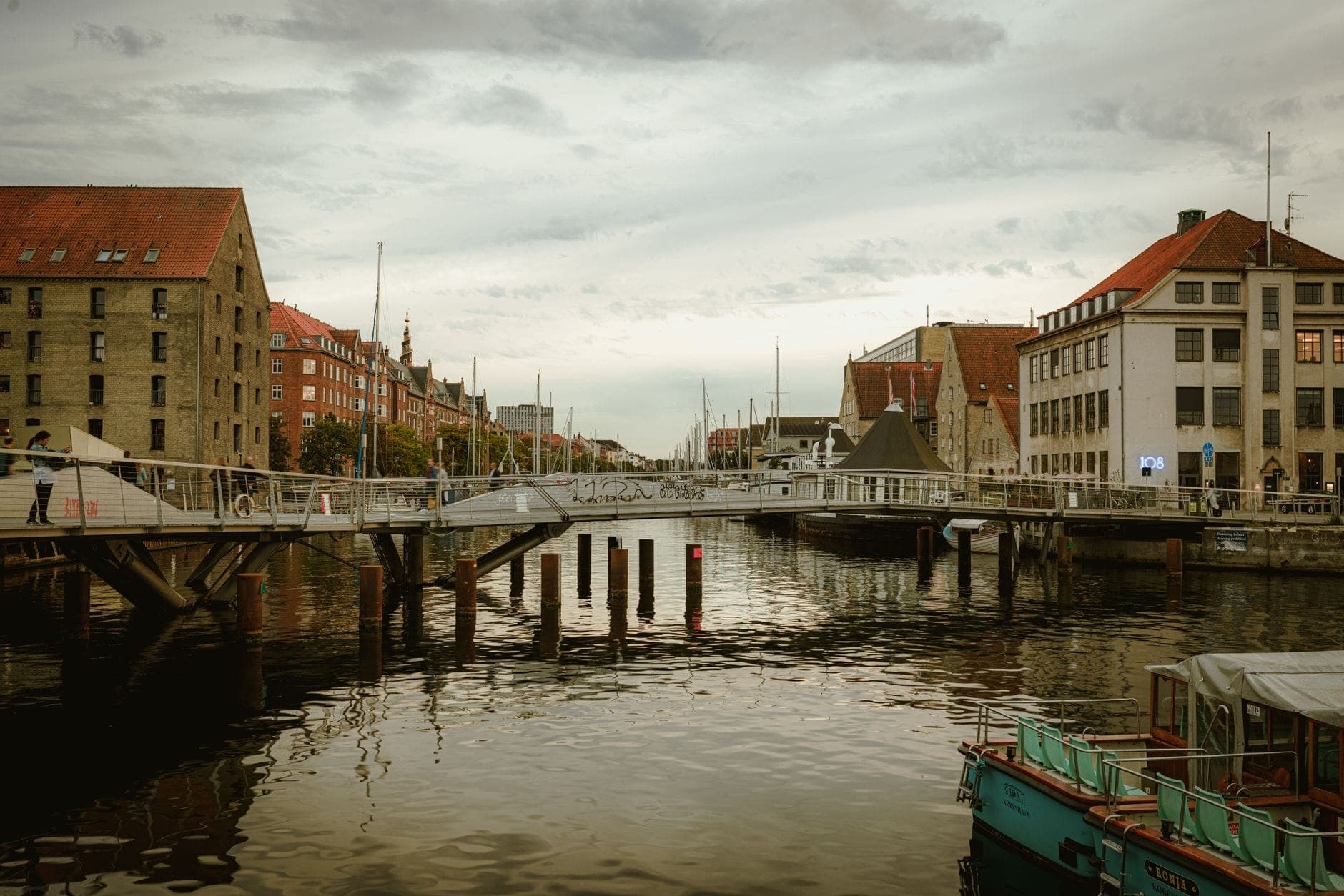 Pemandangan kanal di Copenhagen dengan jembatan pejalan kaki modern, bangunan bersejarah beratap merah, perahu, dan lanskap kota di bawah langit berawan.
