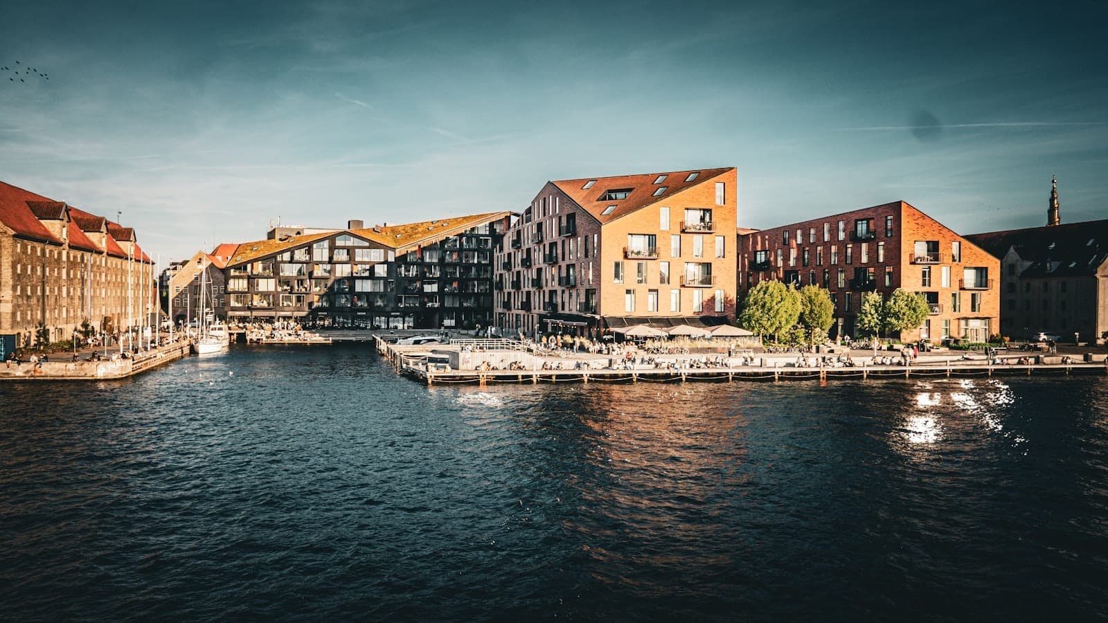 Bangunan tepi laut modern di Kopenhagen saat golden hour, menampilkan arsitektur khas Denmark yang terpantul di air dan suasana hangat yang hidup.