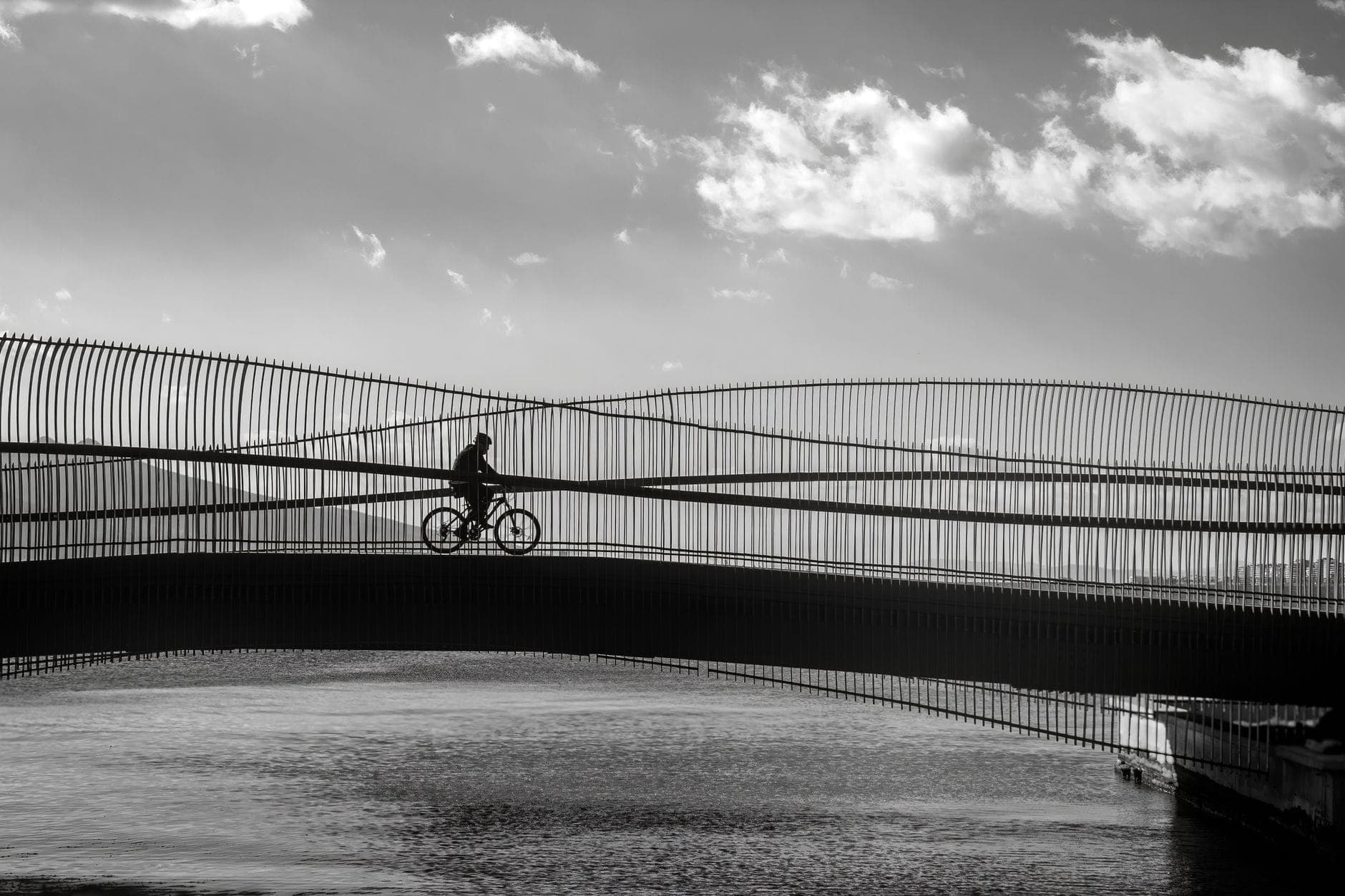 Siluet seorang pengendara sepeda yang melintasi jembatan modern dengan desain arsitektur khas di atas air di Kopenhagen saat matahari terbenam.