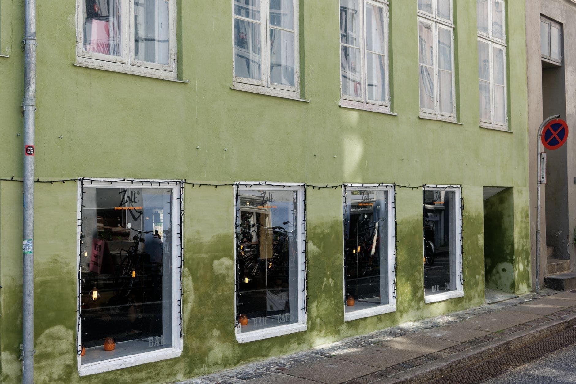 Exterior de um restaurante moderno em Copenhague com fachada pintada de verde e grandes janelas, refletindo uma elegante iluminação interior escandinava.