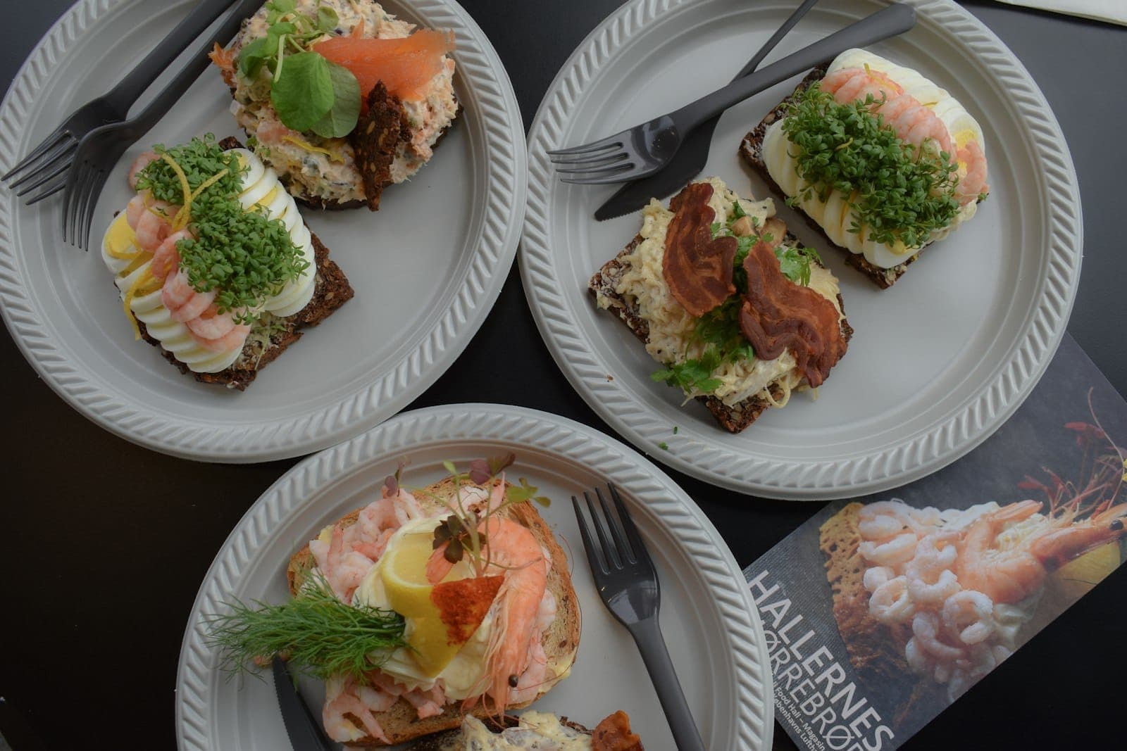 Vier Teller mit traditionellem dänischen Smørrebrød mit verschiedenen Belägen, von oben fotografiert auf einem Tisch – bunt und appetitlich, typisch für Kopenhagens Küche.