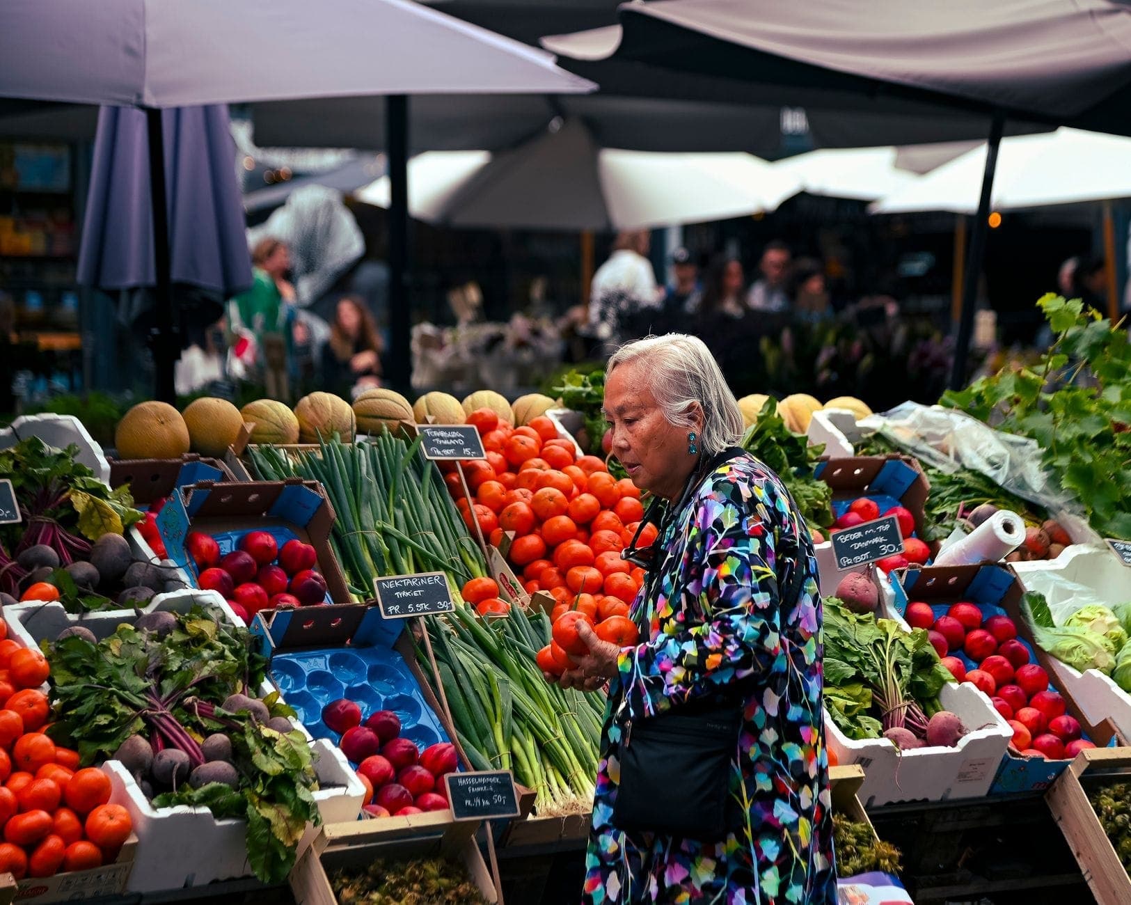 Mulher mais velha fazendo compras em uma barraca de mercado ao ar livre vibrante, cheia de legumes e frutas frescas, com compradores e guarda-chuvas ao fundo.