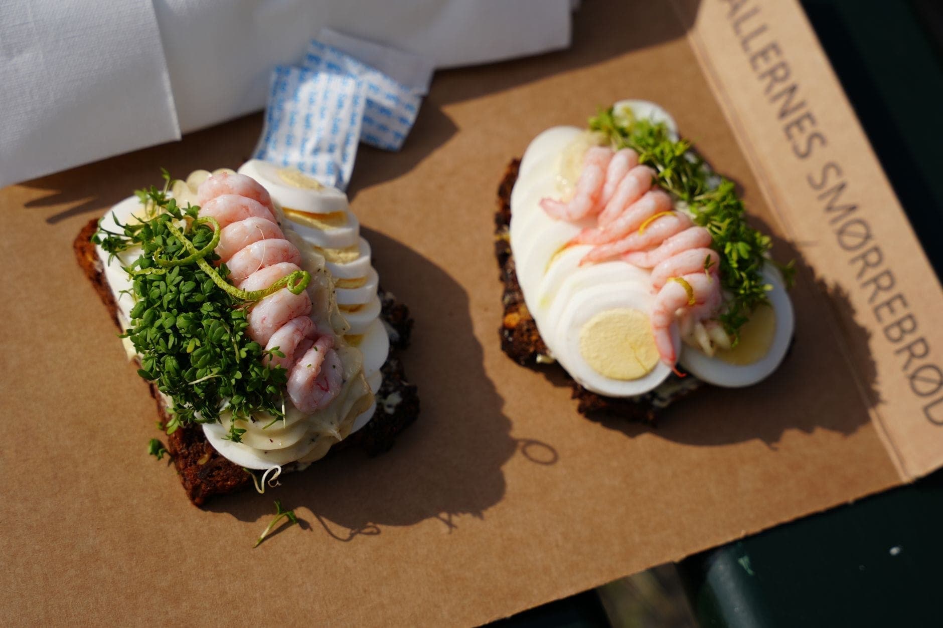 Dois sanduíches abertos dinamarqueses, smørrebrød, com camarão, ovo e ervas sobre pão de centeio escuro apresentados em uma bandeja de papelão.