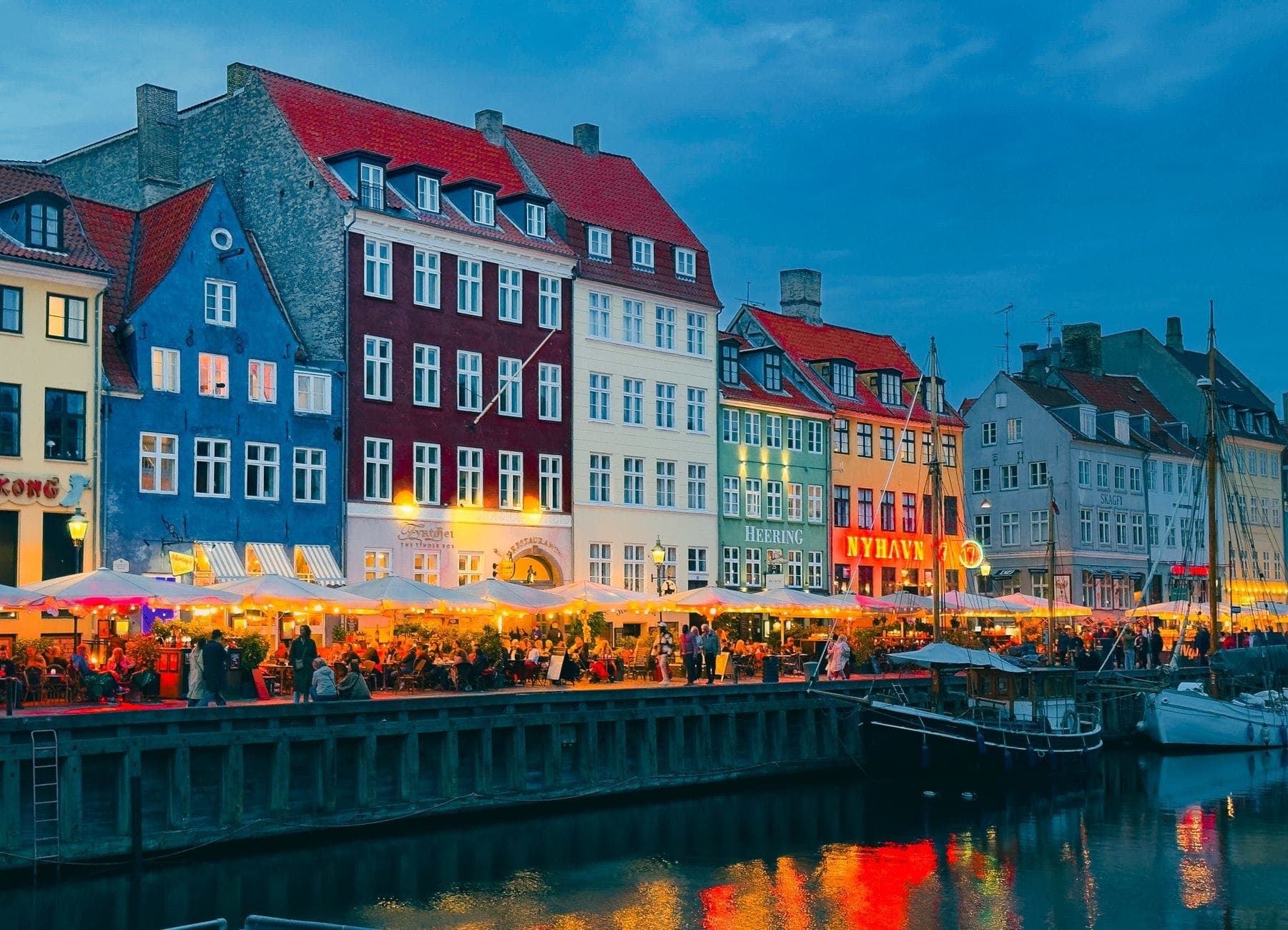 Restaurantes e cafés coloridos e vibrantes à beira do canal em Nyhavn, Copenhague, com clientes jantando à noite e luzes refletidas na água.
