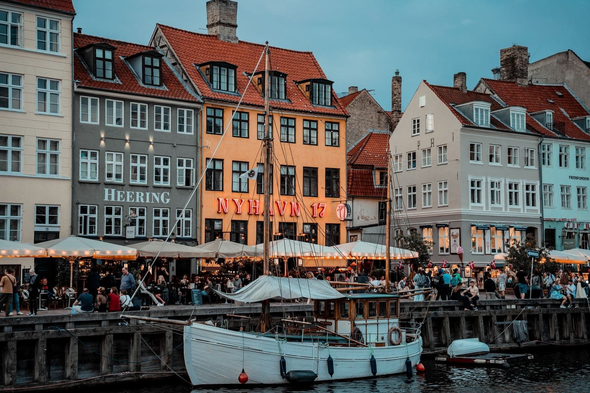Puerto de Nyhavn en Copenhague con restaurantes al aire libre y multitudes cenando por la noche junto a coloridos edificios históricos.