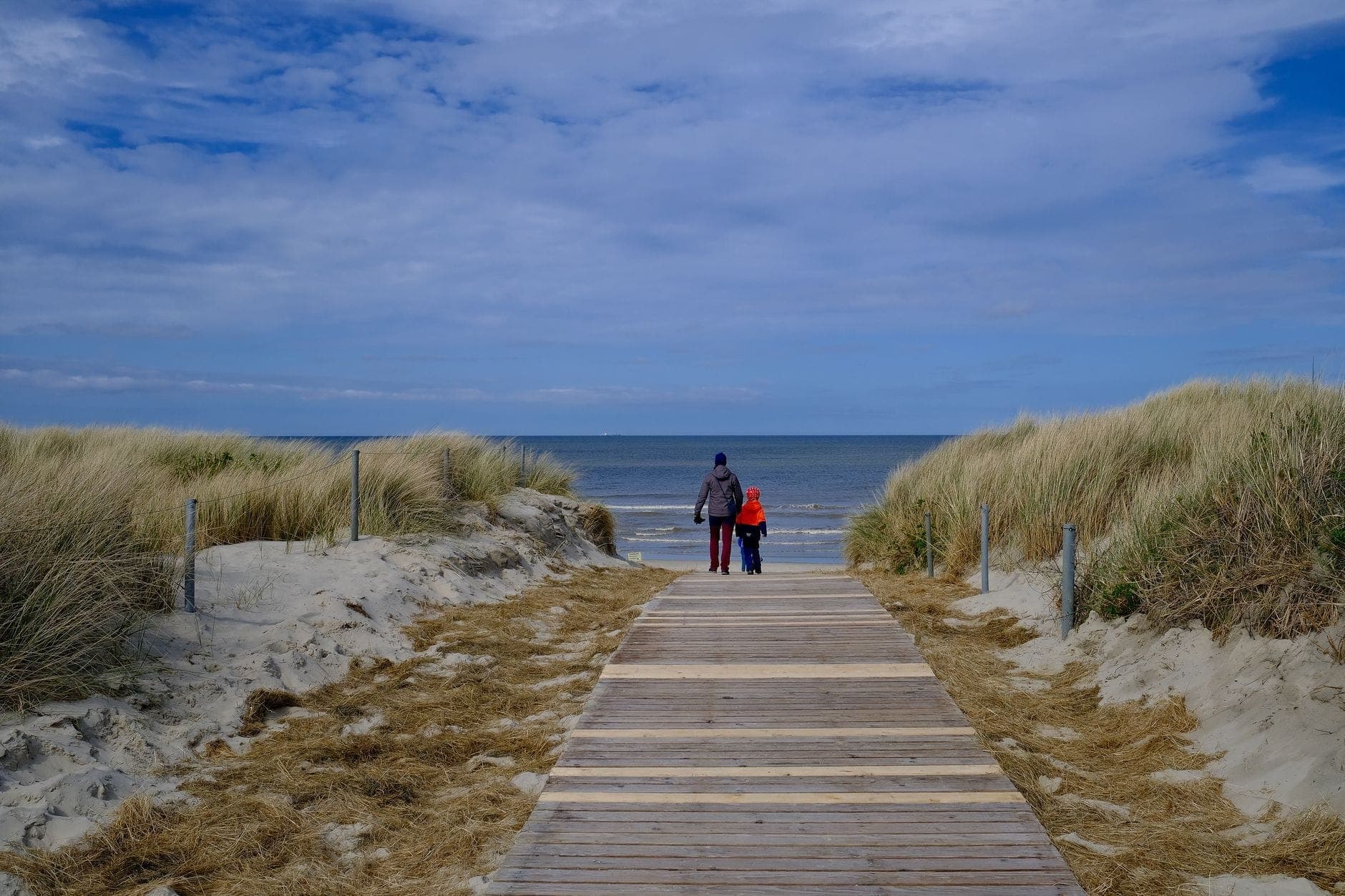 Dos personas con chaquetas caminan hacia una playa desierta por un paseo entablado, con cielo nublado y orilla fría y vacía.