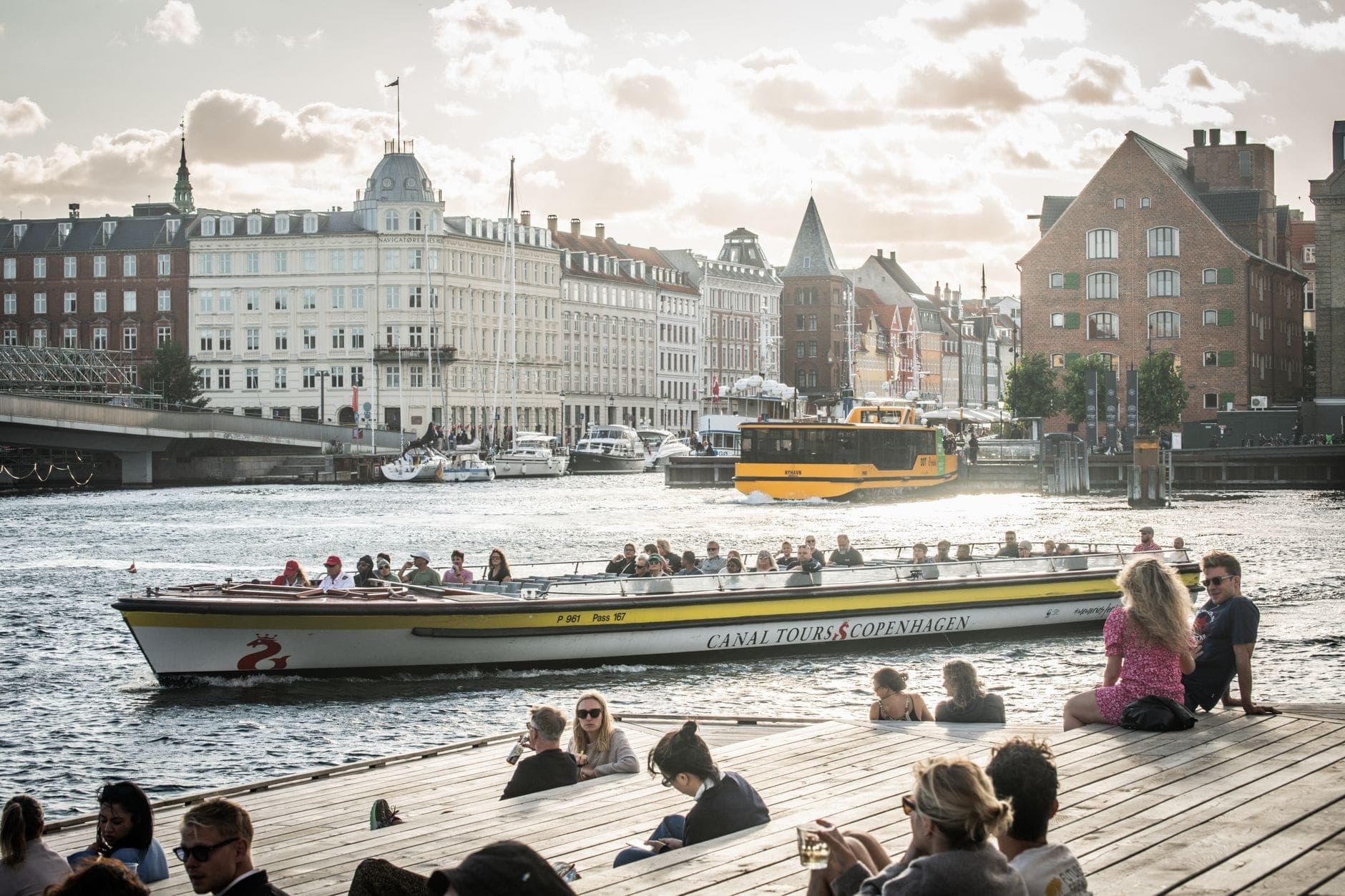 Menschen entspannen auf einem Holzsteg in Kopenhagen und beobachten Kanalboote auf dem Wasser, im Hintergrund historische Gebäude an einem sonnigen Sommertag.