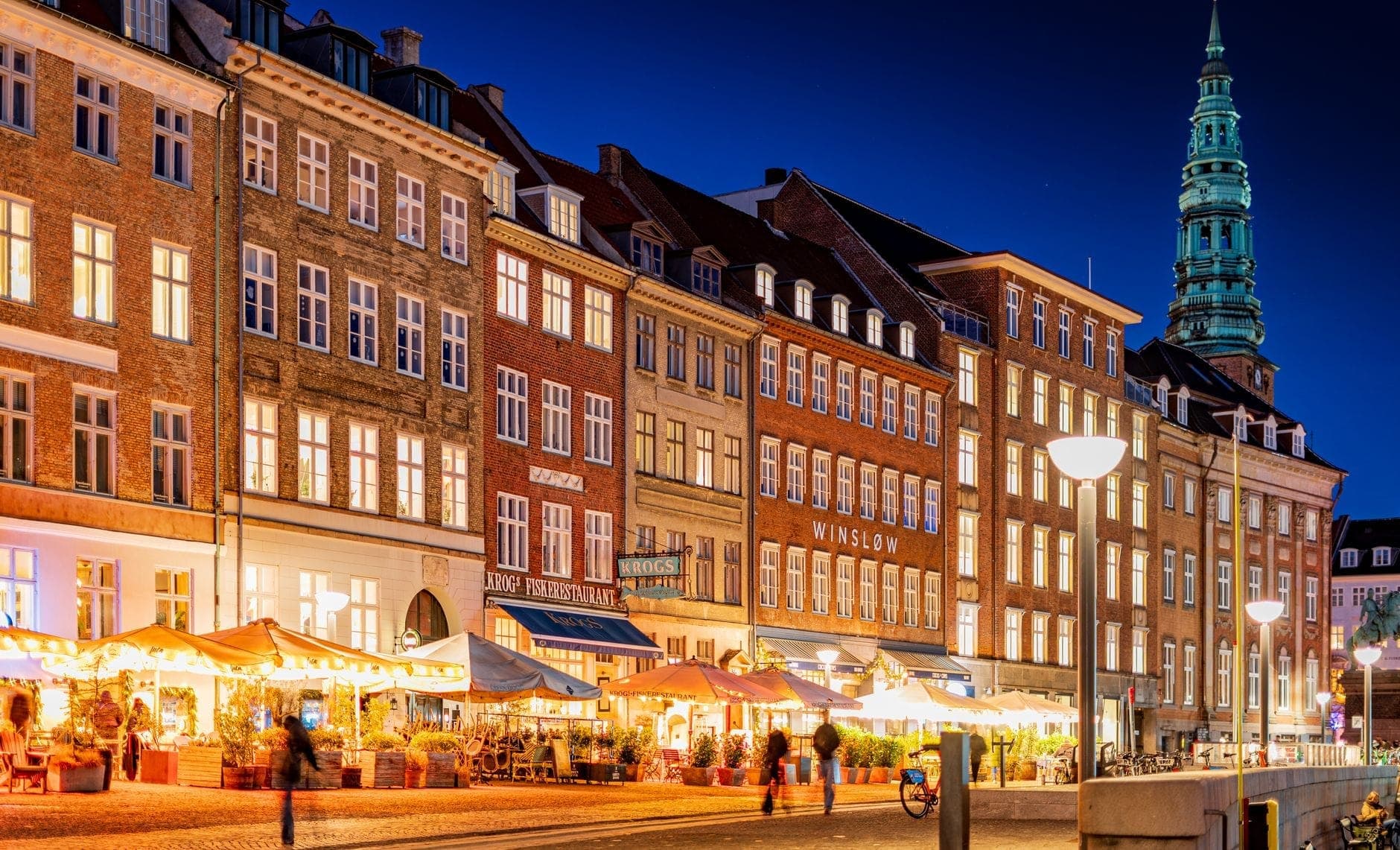 Nächtlicher Blick auf eine belebte Kopenhagener Straße mit lebhaften Bars und Restaurants, bunten historischen Gebäuden und Menschen, die das lebendige Nachtleben genießen.