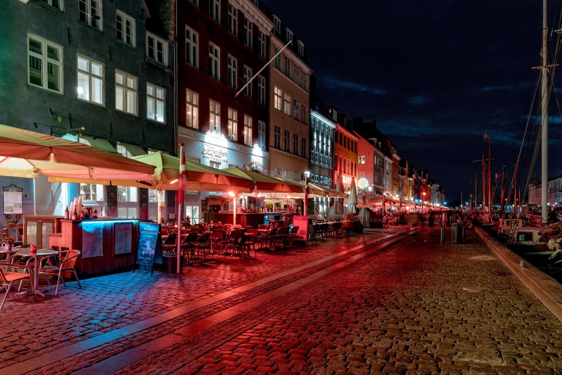 Vista noturna de Nyhavn em Copenhague com bares e restaurantes iluminados ao longo do canal, áreas de estar ao ar livre e iluminação urbana atmosférica.