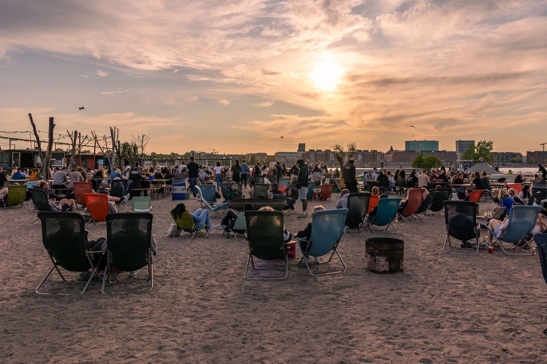 Pessoas relaxando em cadeiras de praia coloridas na areia ao pôr do sol perto da orla de Copenhagen, aproveitando uma animada cena de verão ao ar livre.
