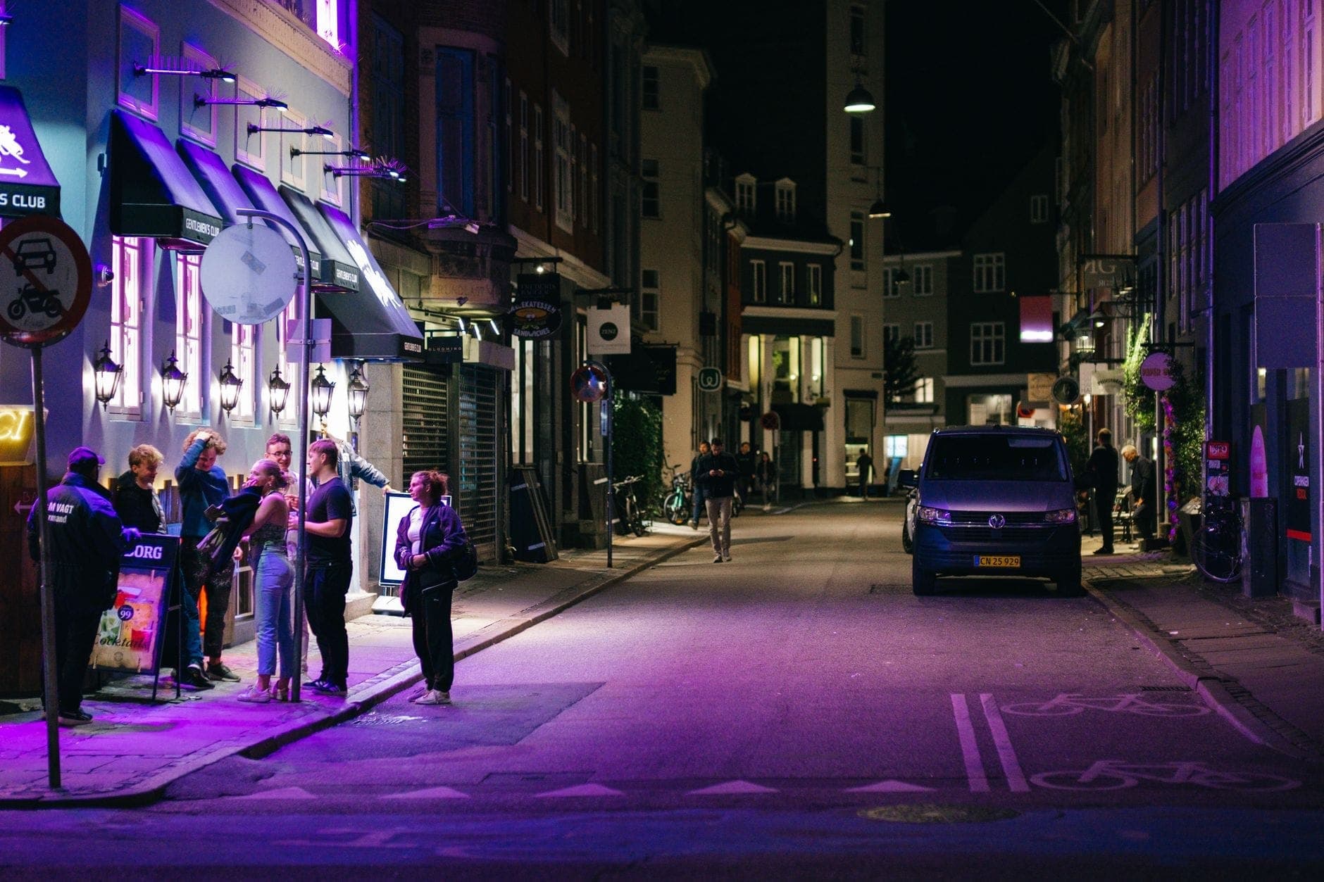 Pessoas em pé e socializando em uma rua animada iluminada por néons em frente a bares em Copenhague à noite.