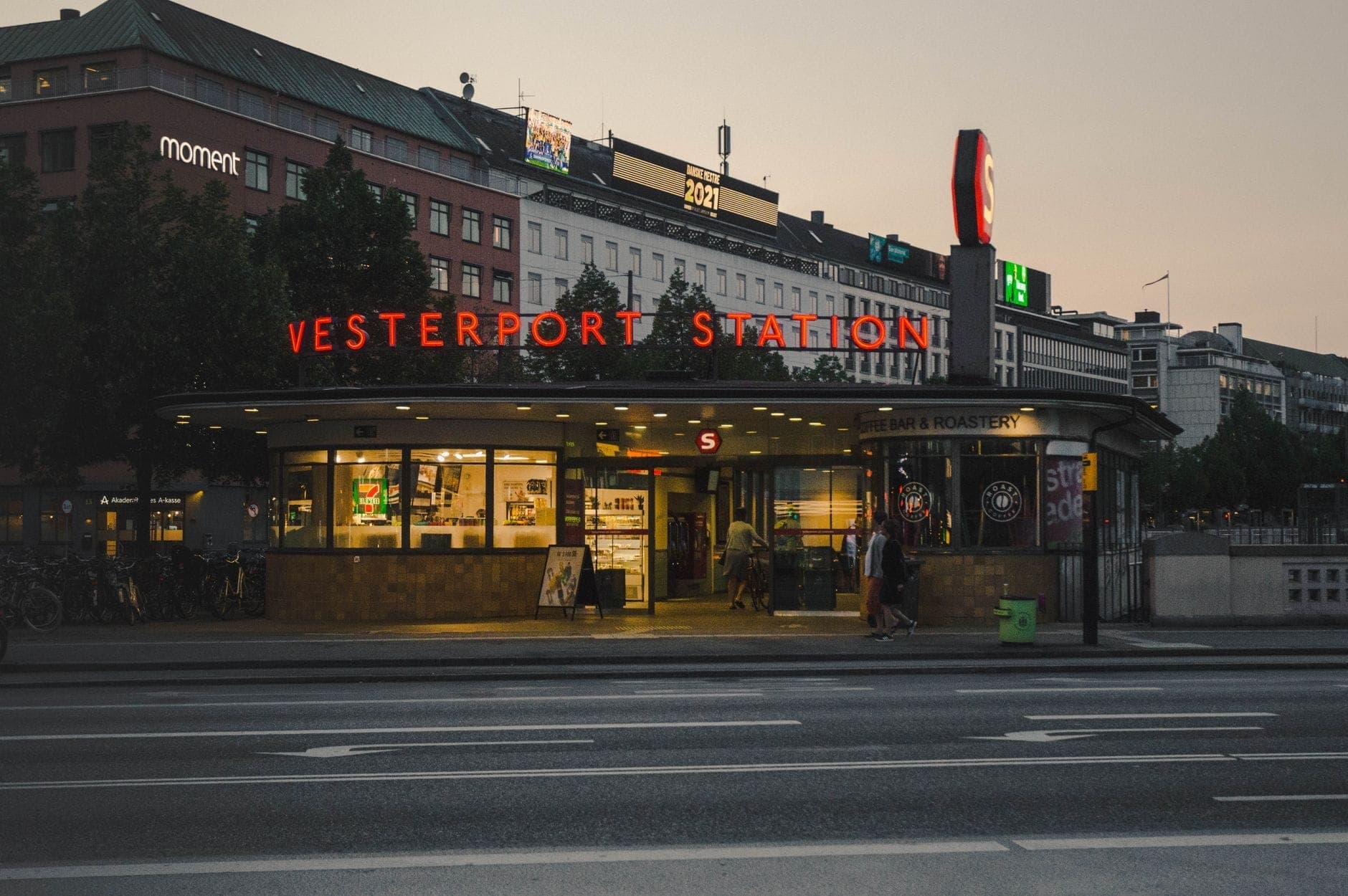 Entrada da estação Vesterport ao anoitecer em Copenhague com sinalização iluminada e edifícios da cidade ao fundo.