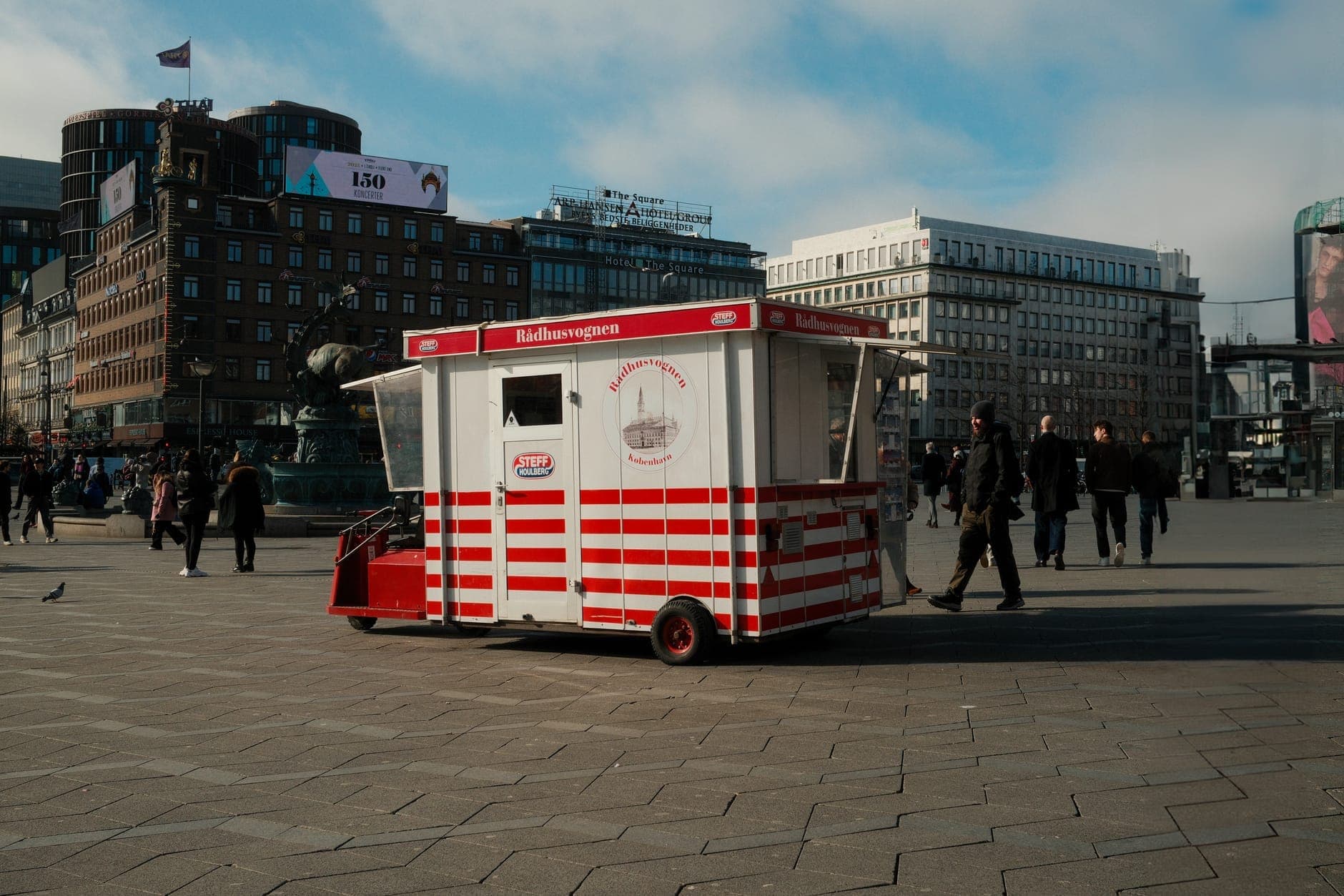 Clásico pølsevogn (carrito de perritos calientes) danés rojo y blanco en una concurrida plaza de la ciudad de Copenhague, con peatones y edificios urbanos al fondo.