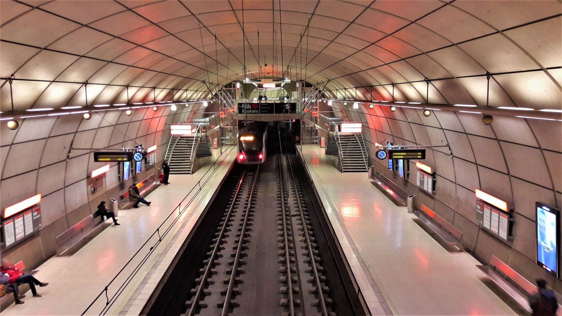 Estación de metro subterránea moderna en Copenhague con un tren llegando, personas esperando en el andén y señales iluminadas.