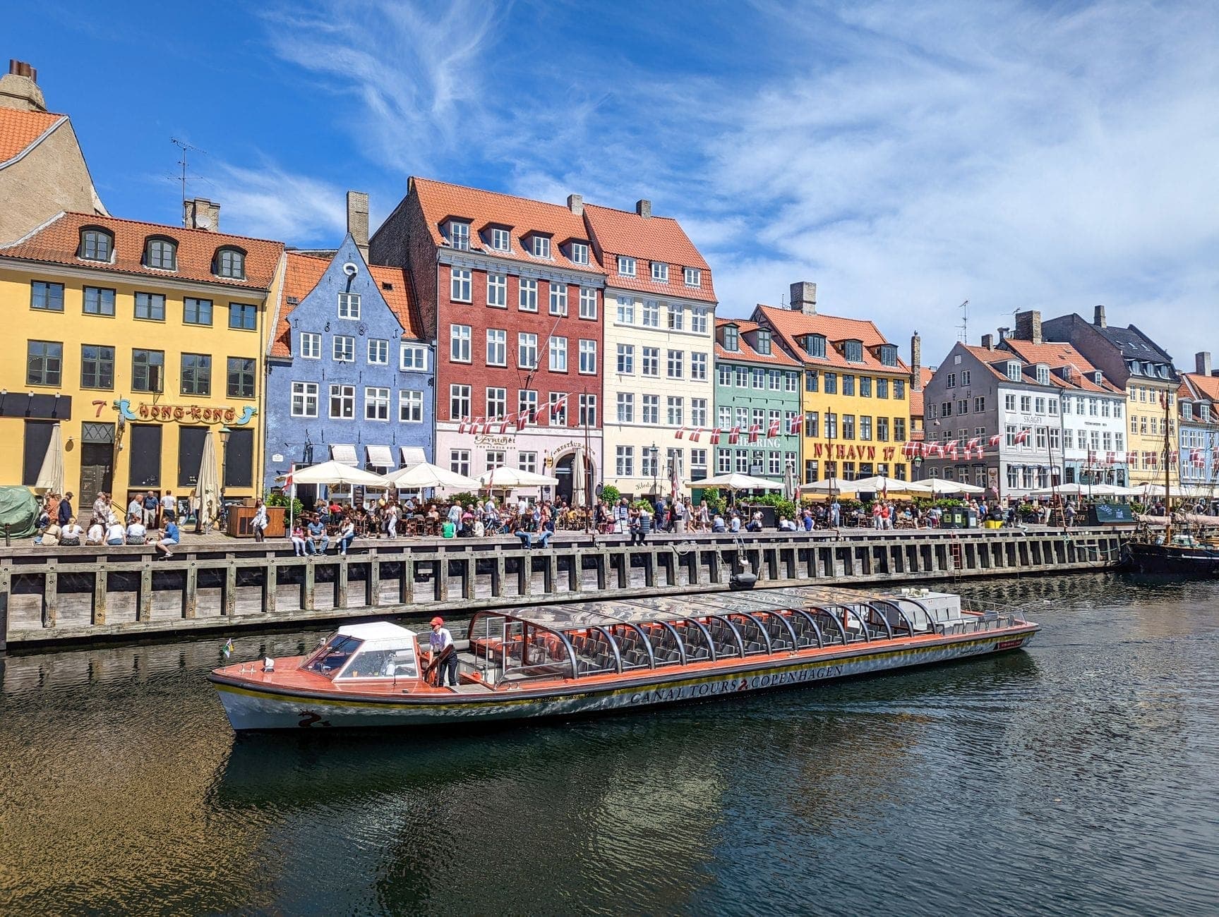 Bunte historische Häuser säumen das Nyhavn-Kanalufer in Kopenhagen an einem sonnigen Tag, während ein Ausflugsboot vorbeifährt und Menschen in Straßencafés sitzen.