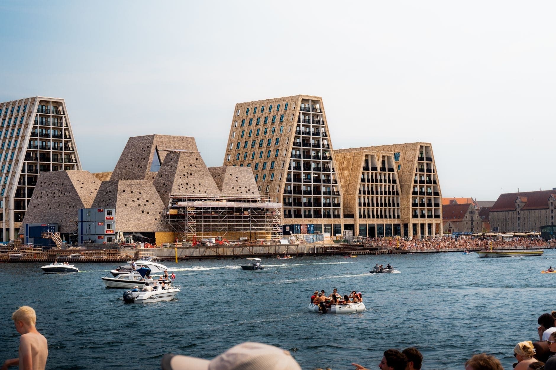 Belebter Sommertag an den Hafenbädern von Copenhagen mit Menschenmengen am Ufer, Booten und Schwimmern, die das Wasser genießen, und markanten modernen Gebäuden im Hintergrund.