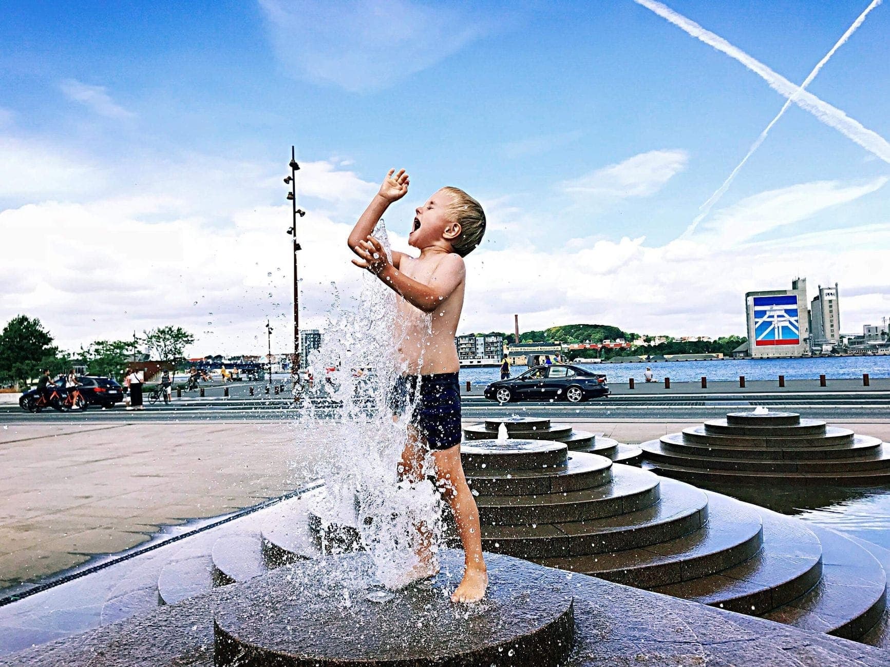Ein kleines Kind plantscht fröhlich in einem Stadtbrunnen, mit Wasserfontänen und einer sonnigen Stadtkulisse im Hintergrund.