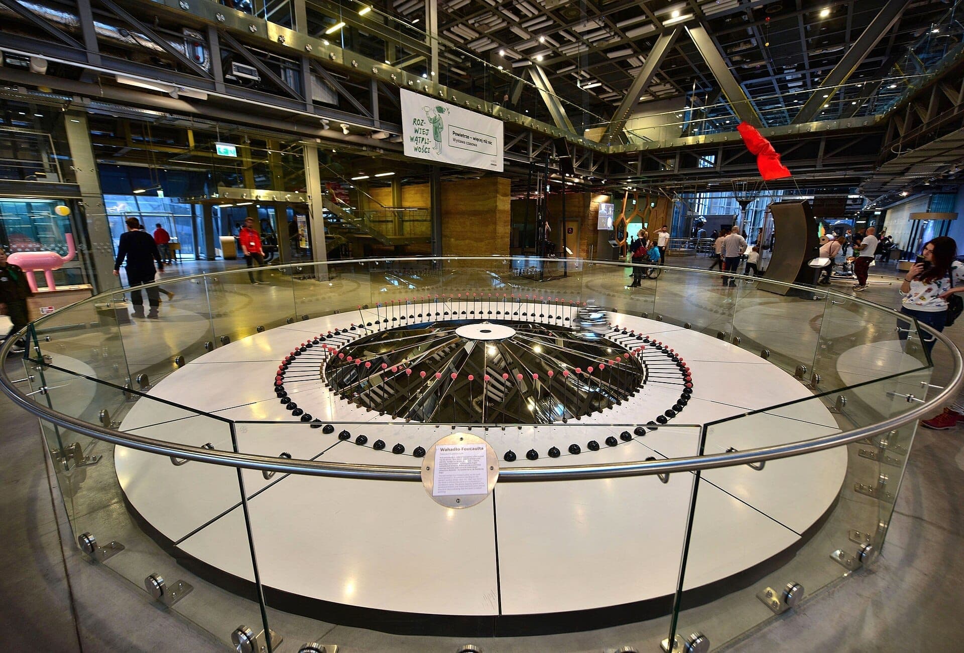 Großes Foucaultsches Pendel, umgeben von einem Glasgeländer im modernen, hell erleuchteten Copernicus Science Centre, mit Besuchern, die den Raum erkunden.