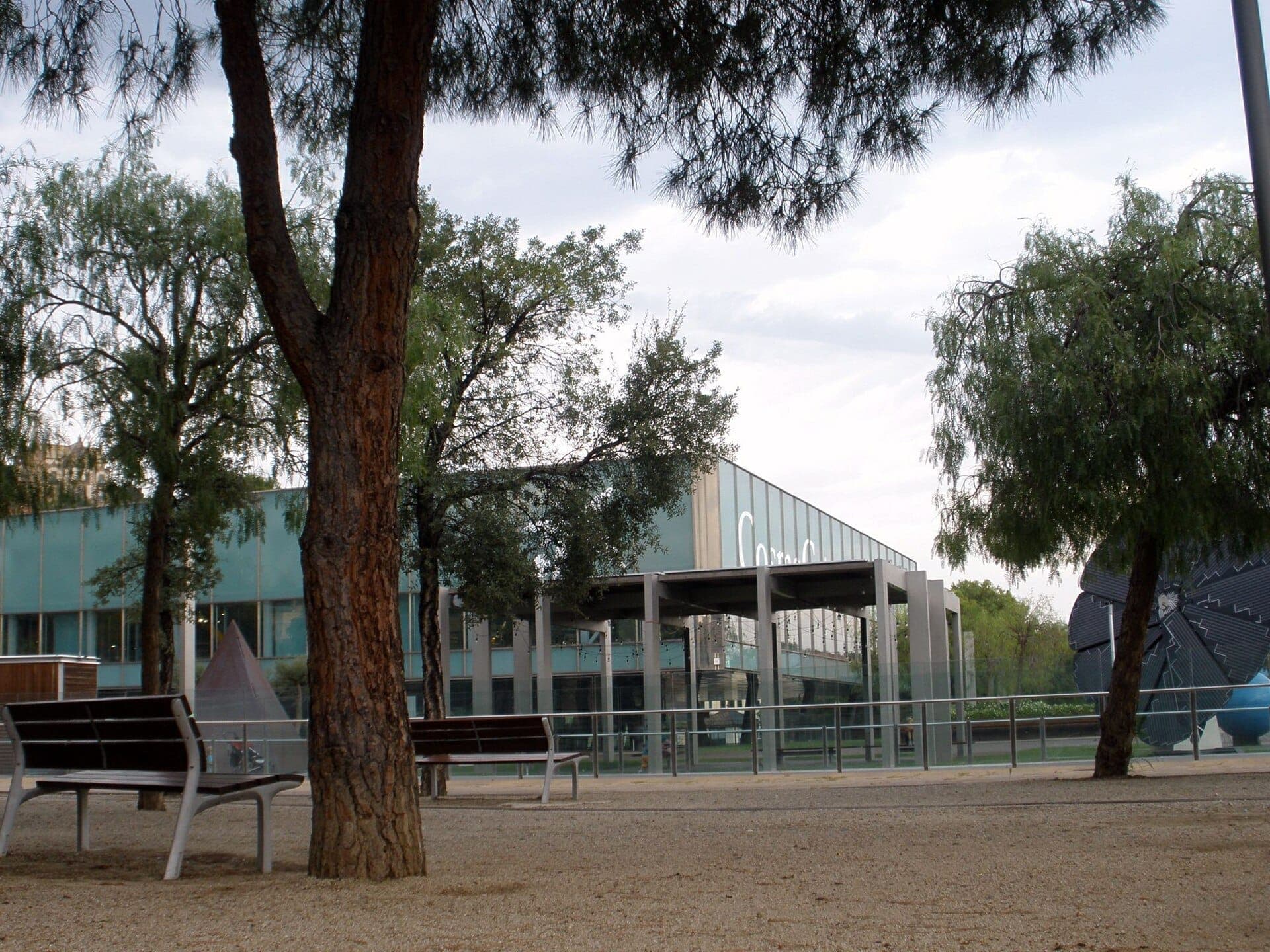 Toàn cảnh bên ngoài CosmoCaixa Barcelona với mặt tiền kính hiện đại, được bao quanh bởi cây xanh và ghế ngồi vào một ngày nhiều mây.