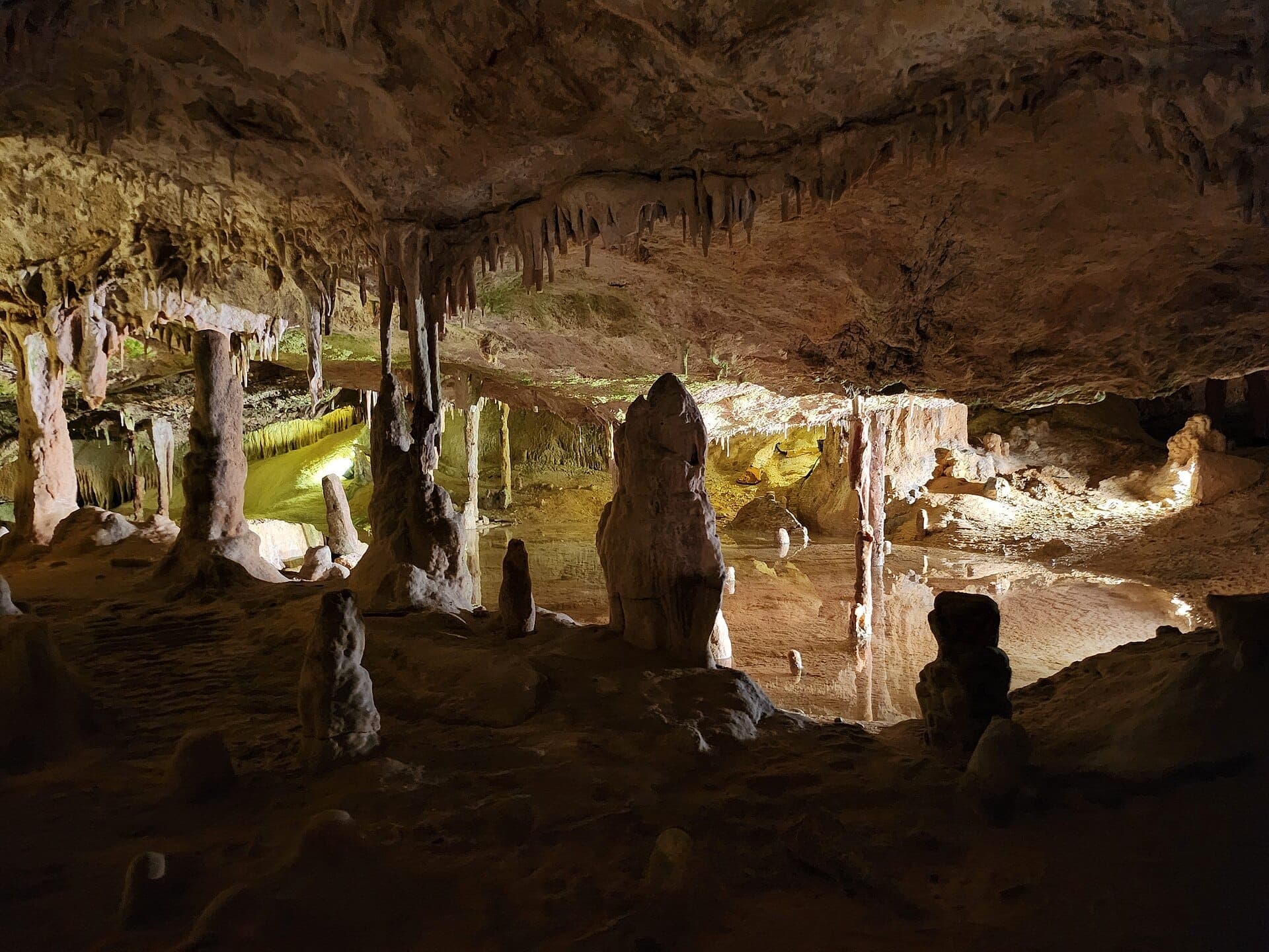 View inside Cova de Can Marçà showing rocky stalactites, tall cave columns, and a reflective underground pool lit by warm lights.