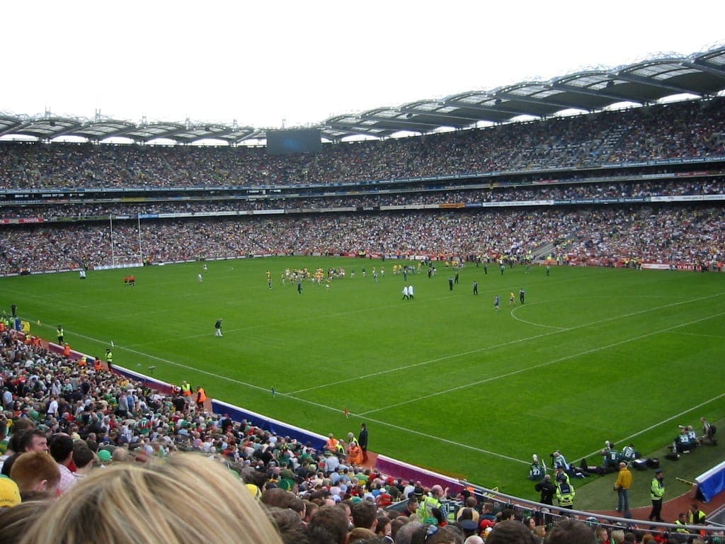 Stadion Croke Park wypełniony tysiącami widzów podczas meczu gier gaelickich, bujnie zielone boisko pod zachmurzonym niebem, tętniąca atmosfera stadionu.