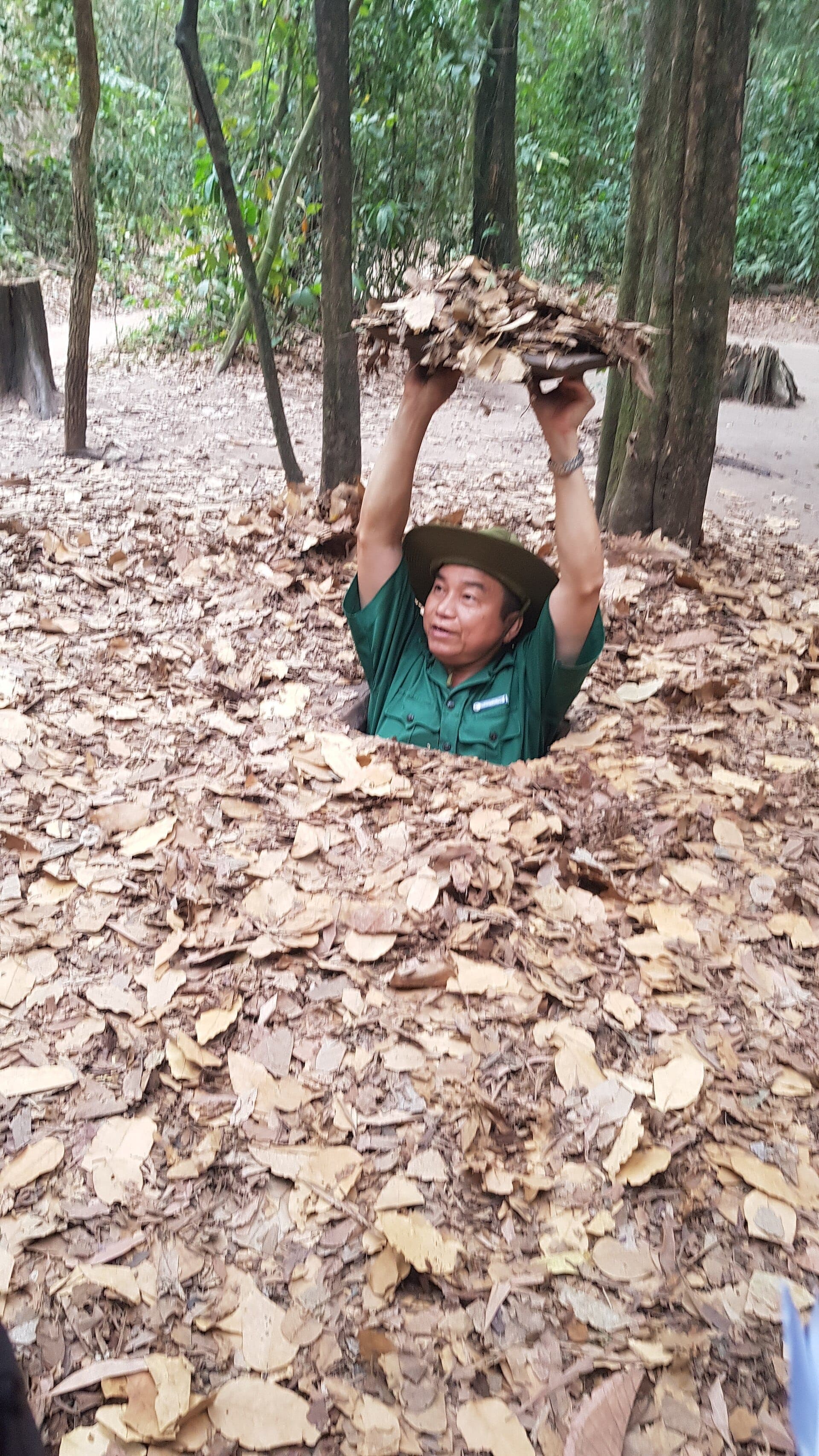 Hướng dẫn viên chui ra từ cửa hầm bí mật phủ đầy lá tại Địa đạo Củ Chi, xung quanh là rừng rậm và mặt đất.
