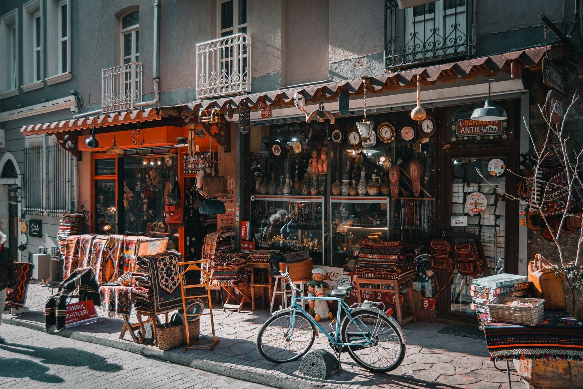 Buntes Antiquitätengeschäft im Antiquitätenviertel Çukurcuma, Istanbul, mit gestapelten Teppichen, Vintage-Gegenständen und einem blaugrünen Fahrrad auf einer Kopfsteinpflasterstraße.