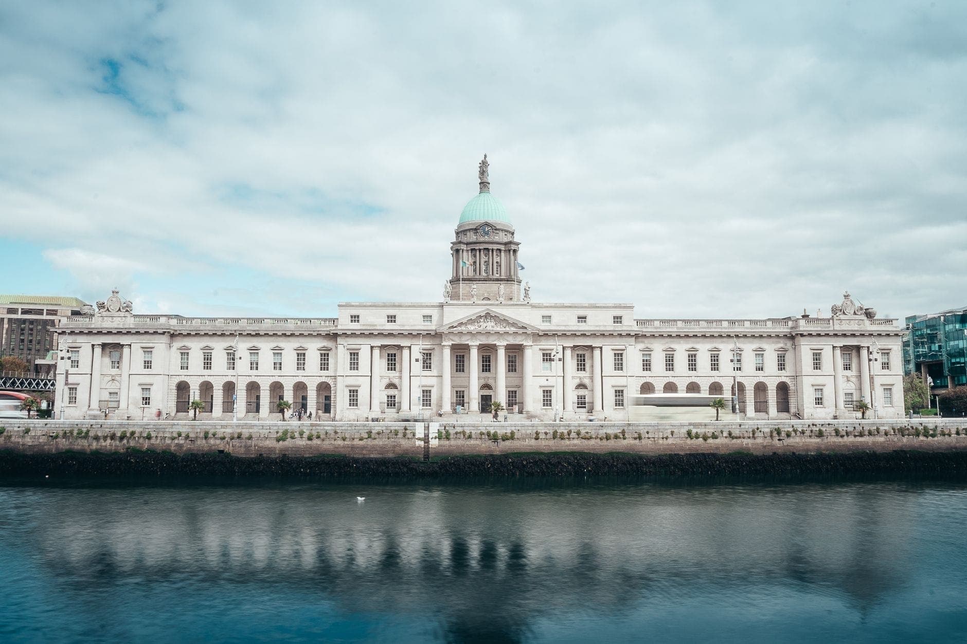 El Custom House de Dublín visto desde la orilla opuesta del río Liffey, con un reflejo nítido en el agua y un cielo dramático con nubes.