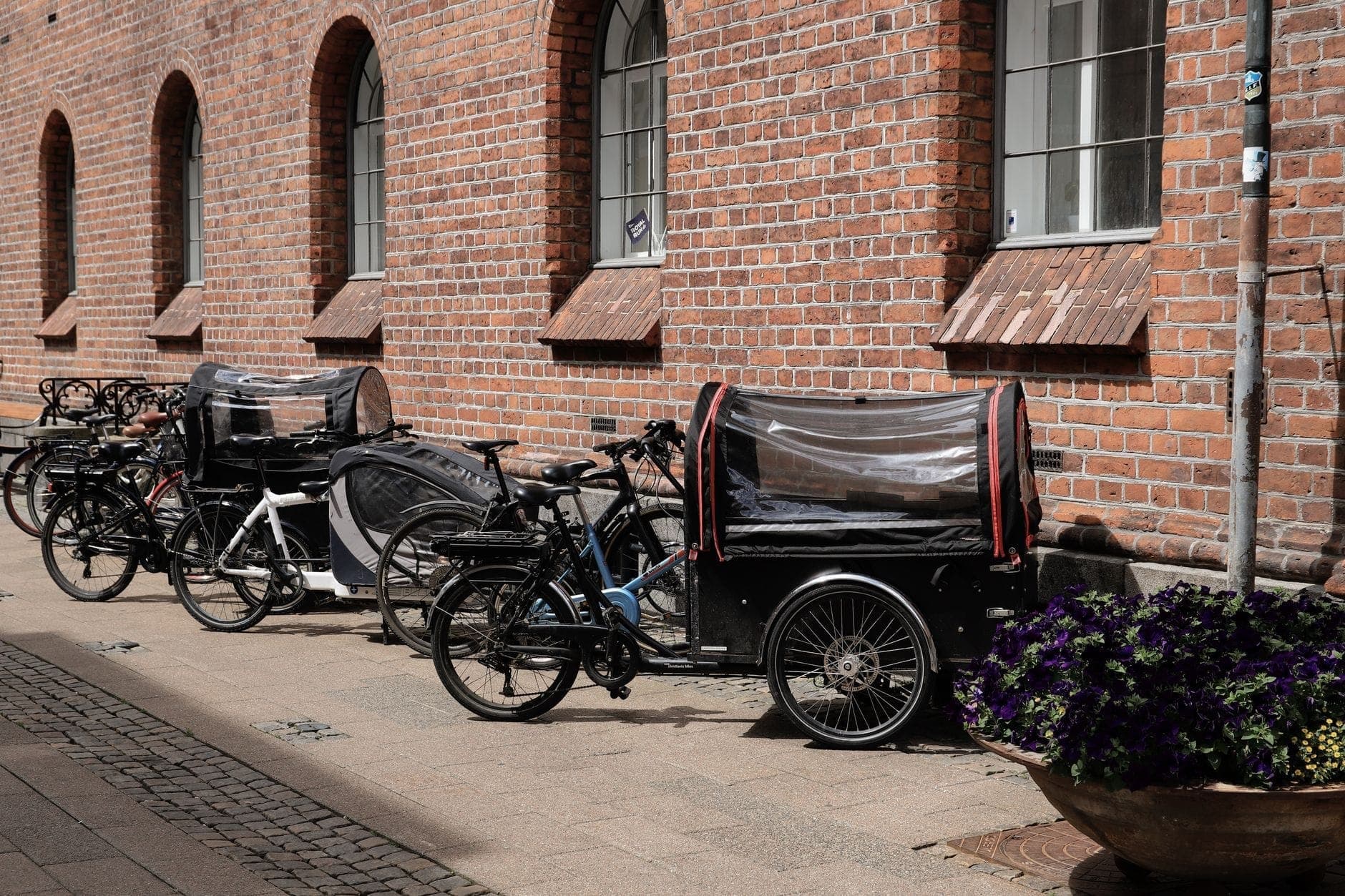 Kilka rowerów, w tym rowery cargo, zaparkowanych jest wzdłuż brukowanej ulicy z czerwoną ceglaną budowlą i łukowymi oknami w Kopenhadze.